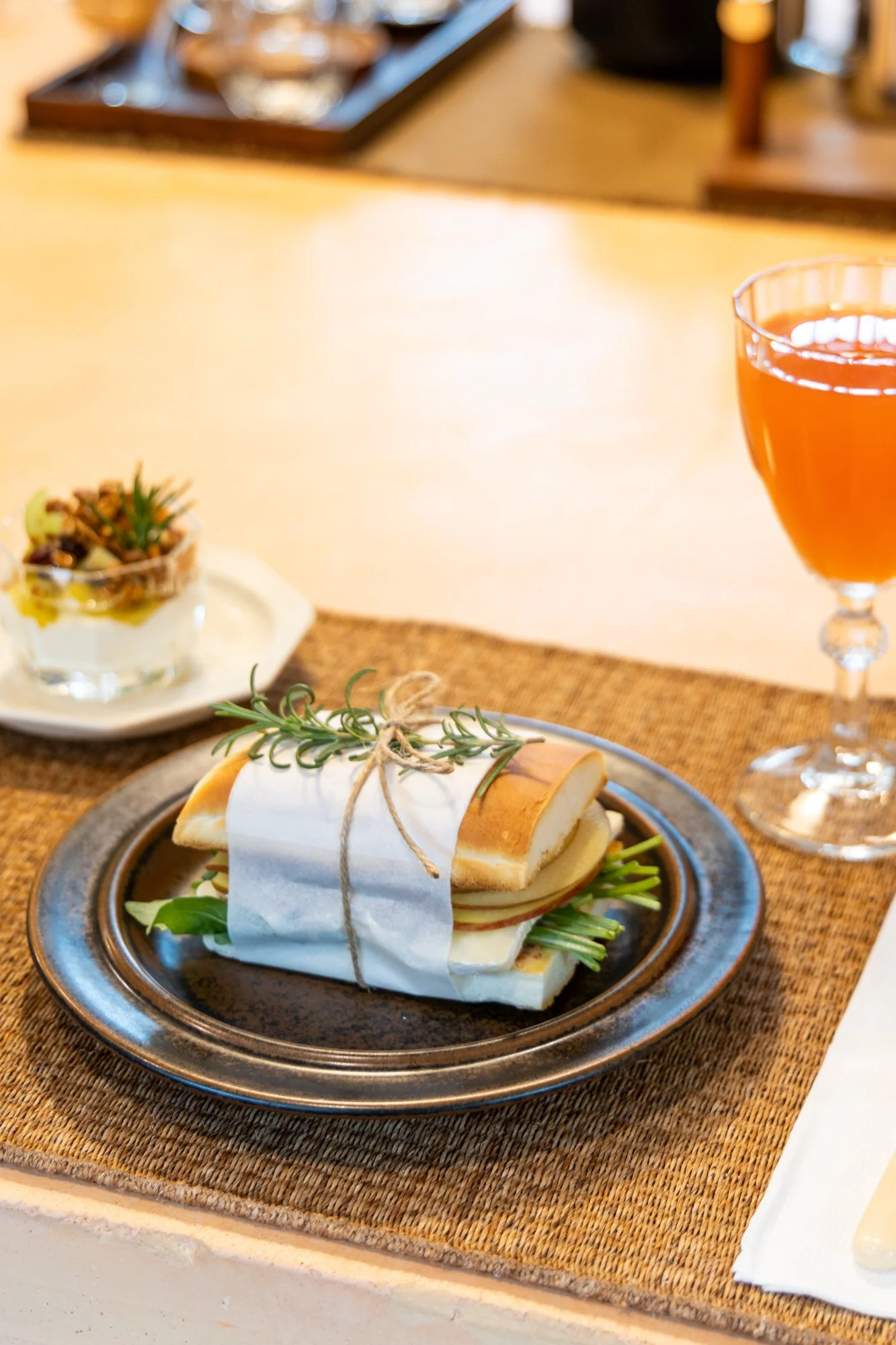 A sandwich wrapped in parchment paper with rosemary sprig on a black plate, a glass of amber-colored beverage, and a small bowl of nuts and dried fruits on a table.