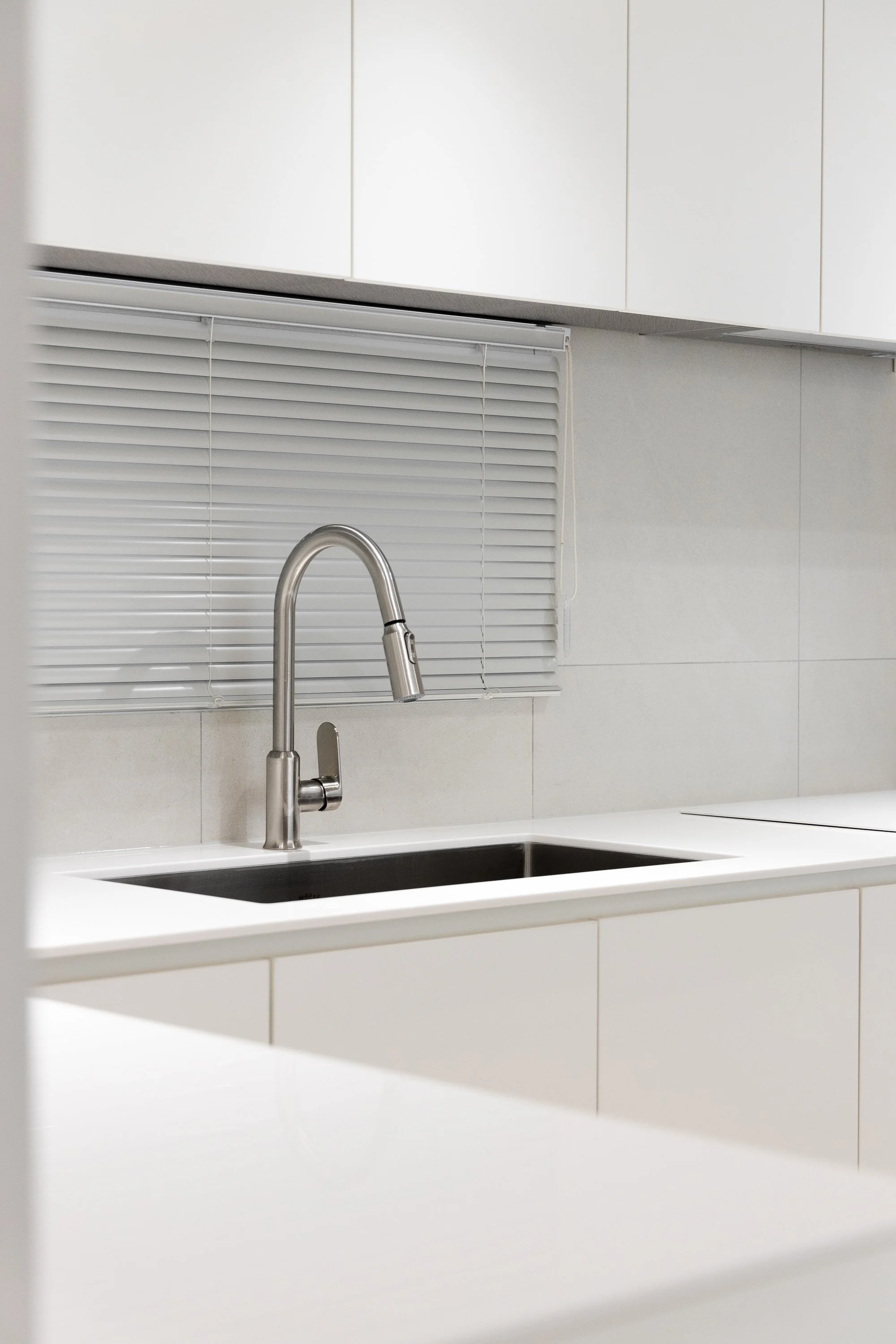 Modern kitchen with a stainless steel faucet, black sink, white countertops, and white cabinets, with a window covered by horizontal blinds.
