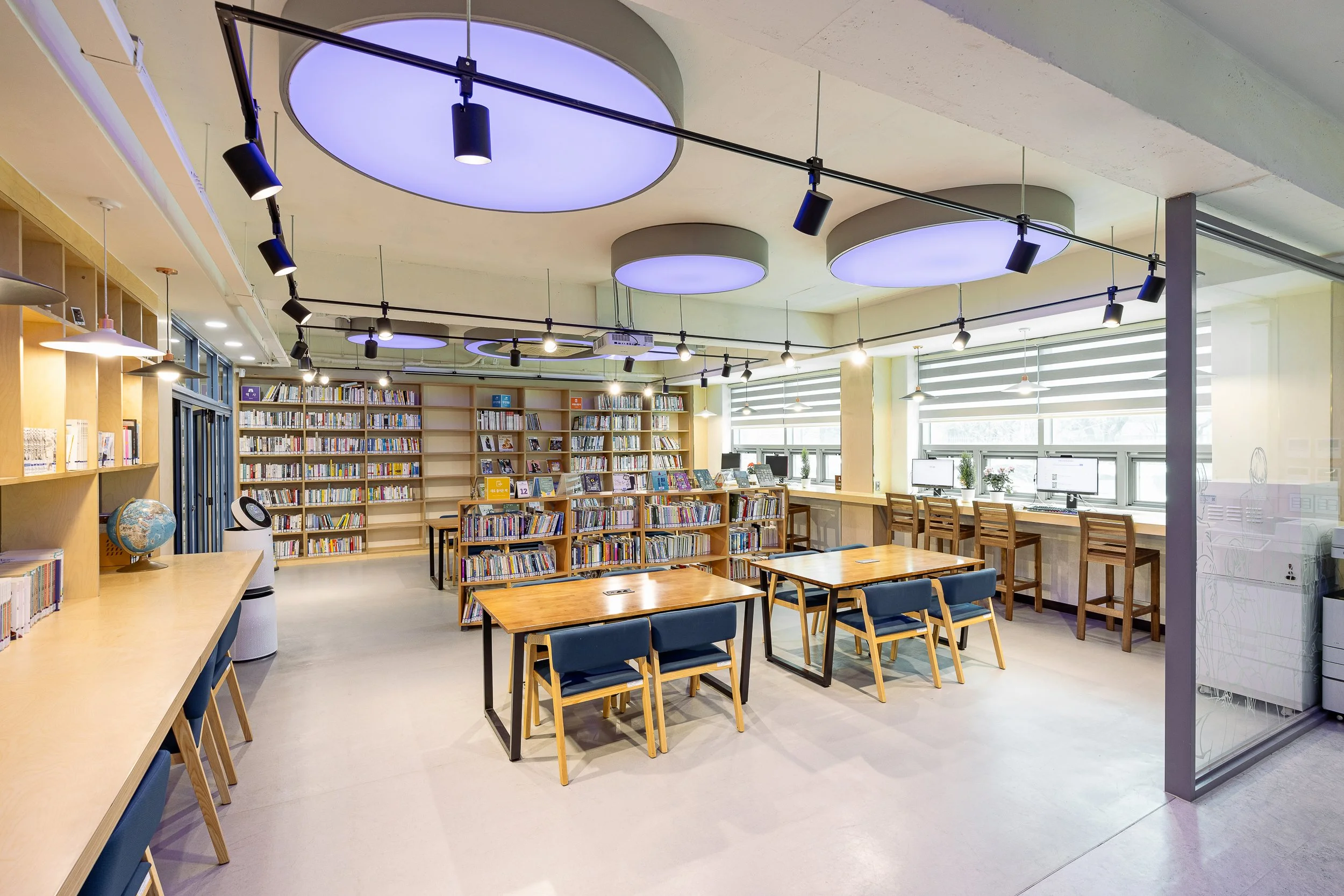 A modern library interior with bookshelves filled with books, wooden tables, and chairs for reading and studying. Large windows with blinds let in natural light, and there are ceiling lights and track lighting on the ceiling.