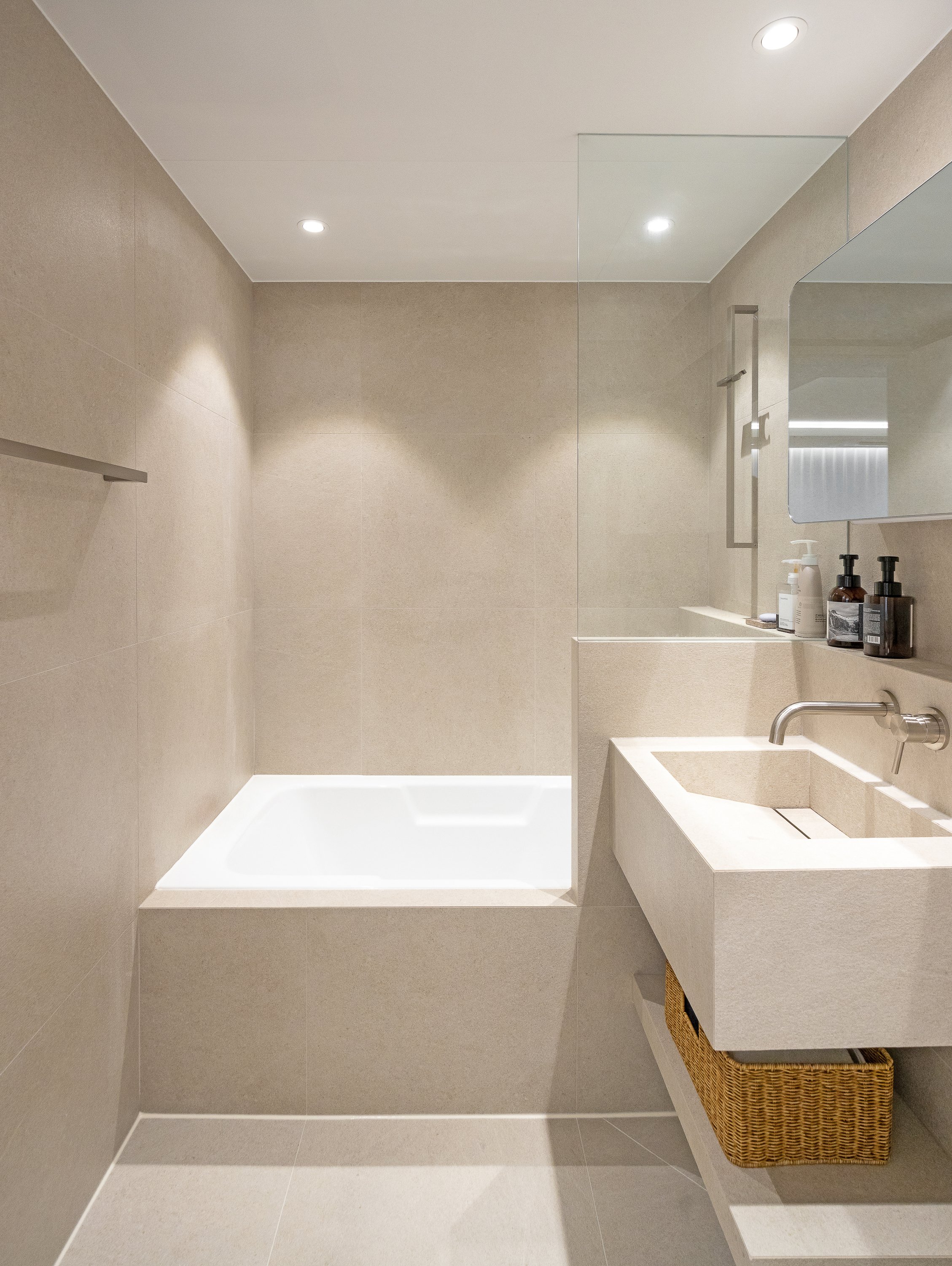Minimalist bathroom with beige tiled walls and floor, a built-in bathtub, a rectangular sink with a wicker basket underneath, and a mirror above the sink with toiletries on the counter.