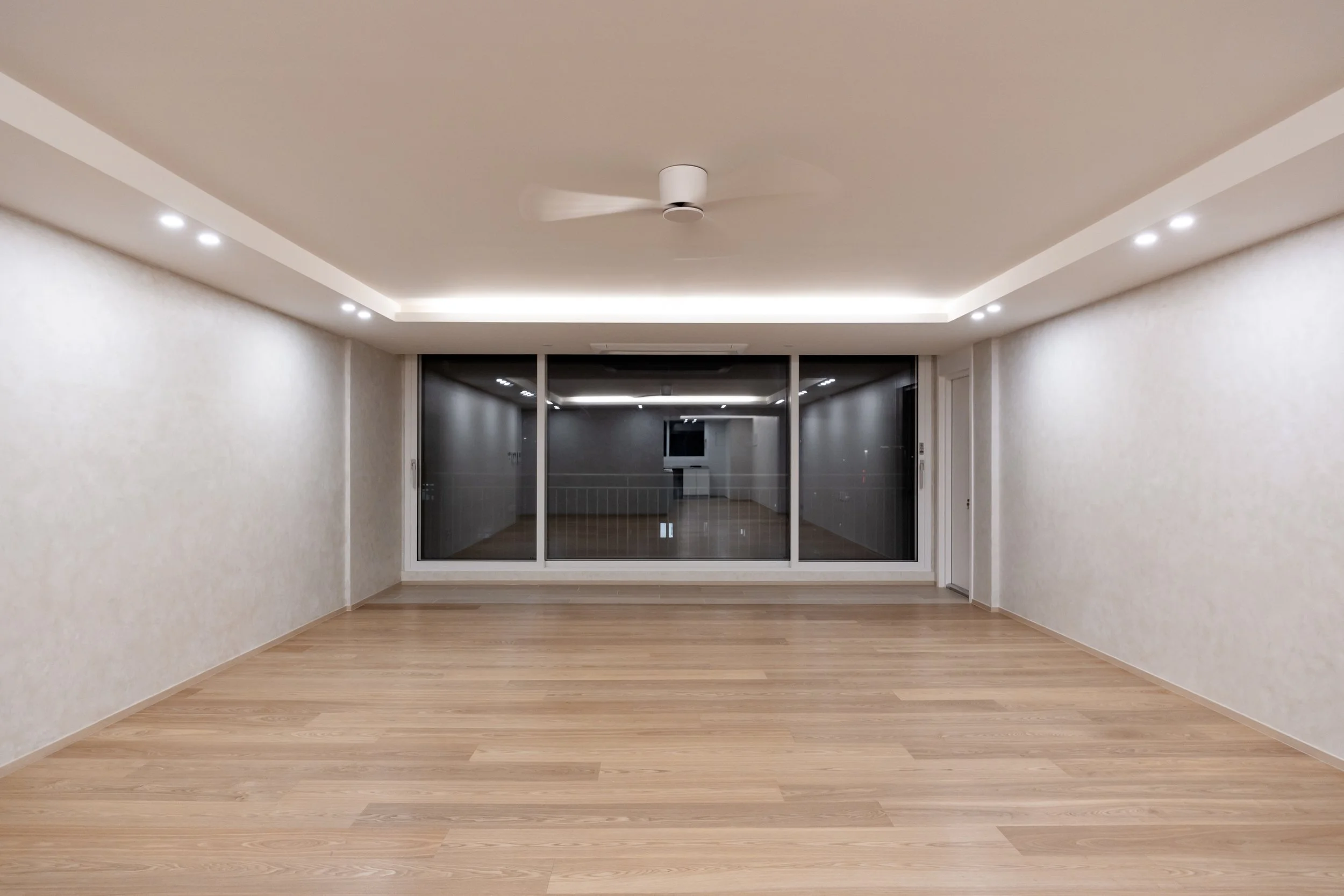 Empty room with wooden floors, white walls, ceiling fan, and large glass sliding doors leading to a balcony or another room.