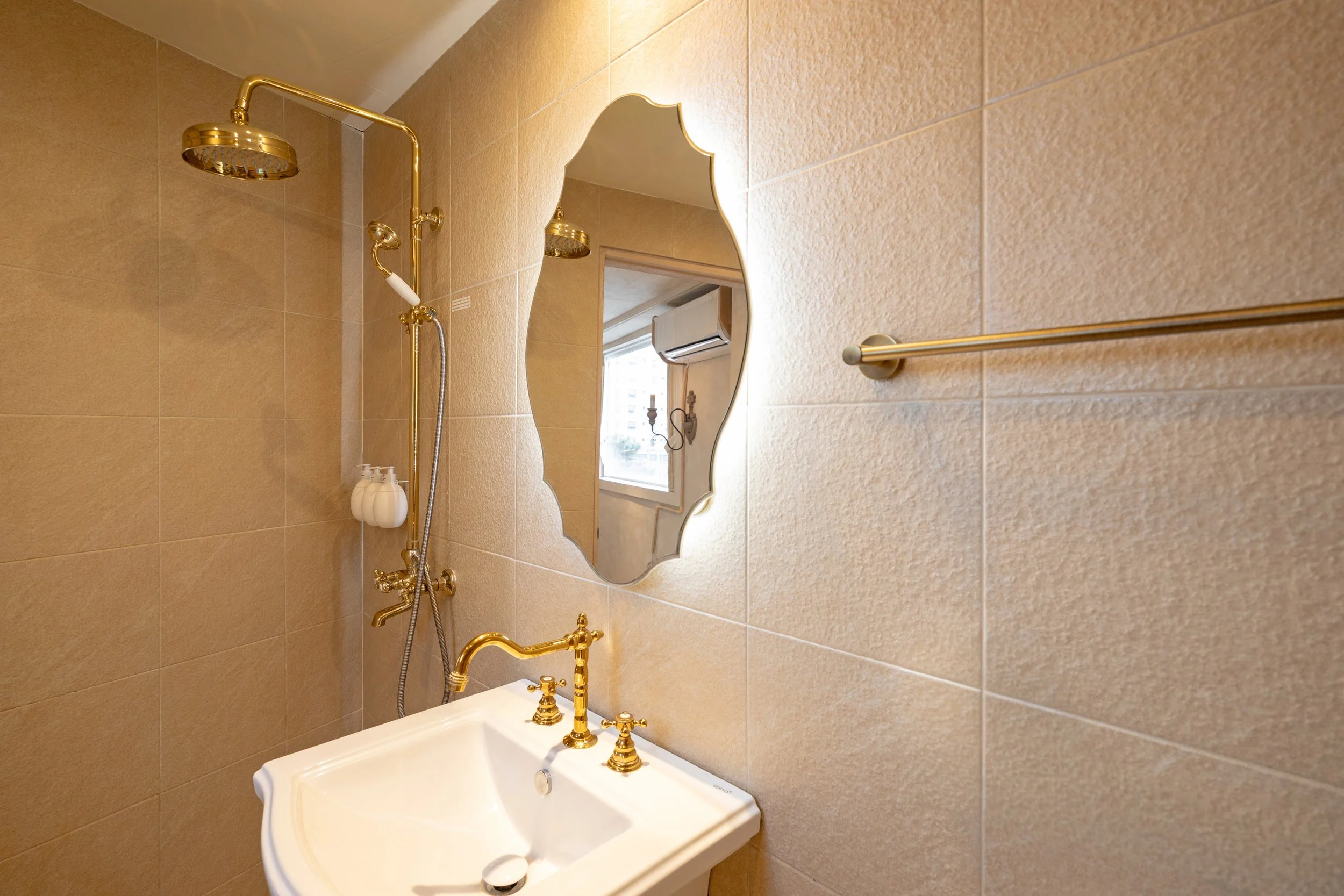 A vintage-style bathroom with beige tiled walls, a white sink with gold faucet and fixtures, a decorative mirror, a gold showerhead, and a window with an air conditioning unit outside.