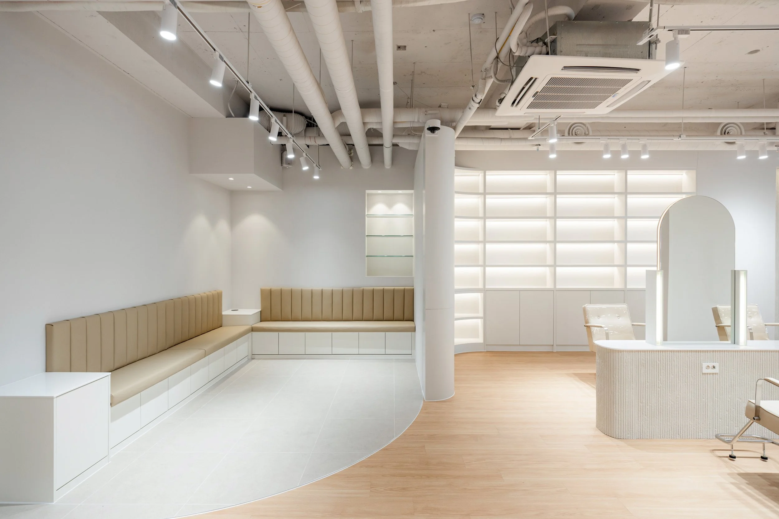 Modern minimalist interior with beige seating, white cabinets, and a curved reception desk, illuminated by track lighting on a white ceiling with visible industrial ducts.