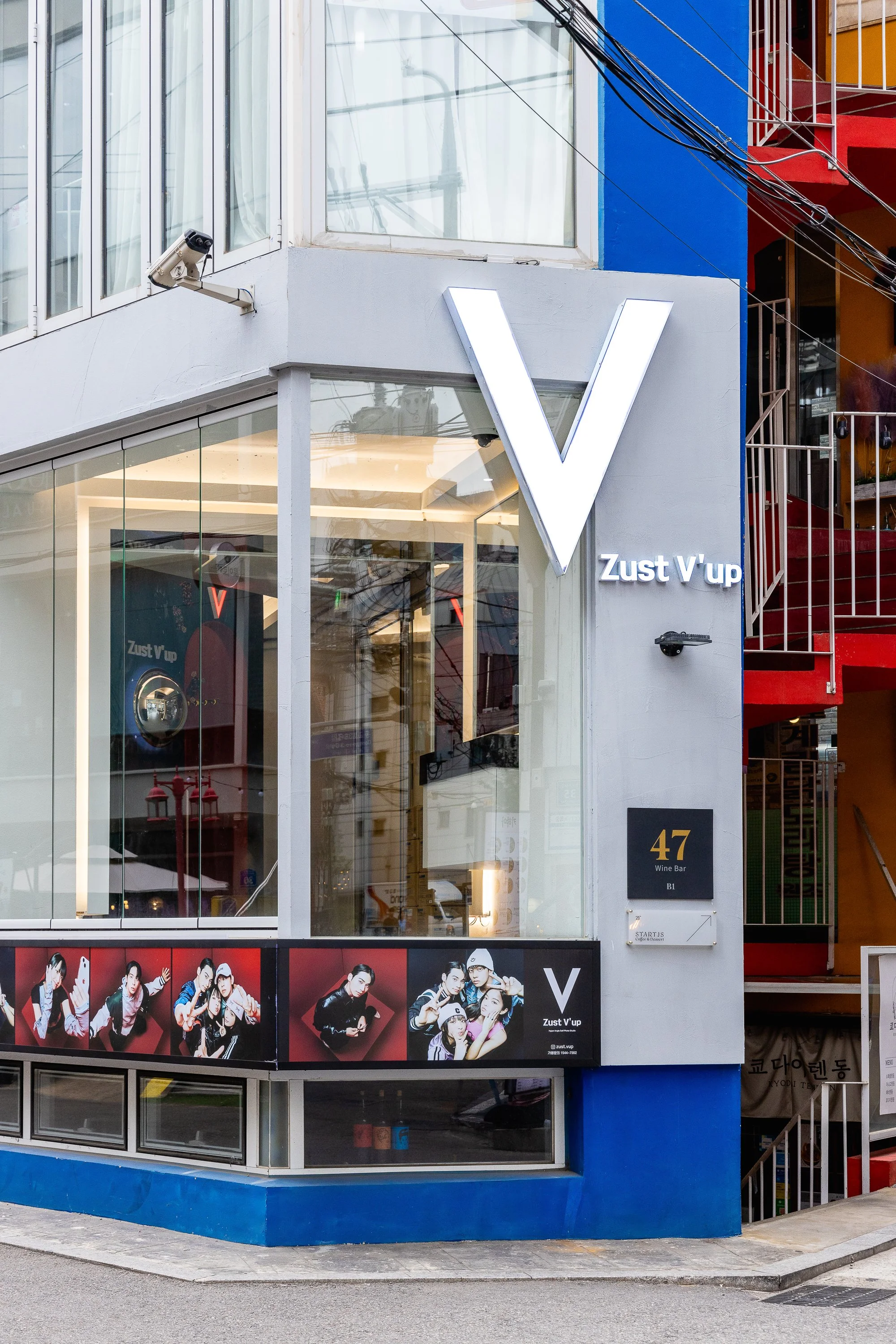 Modern building with large illuminated V sign, advertising 'Zust V'up' nightclub, with a black square sign displaying '47 Wine Bar, B1.' The building has large glass windows, promotional photos of young people, and a security camera installed on the 