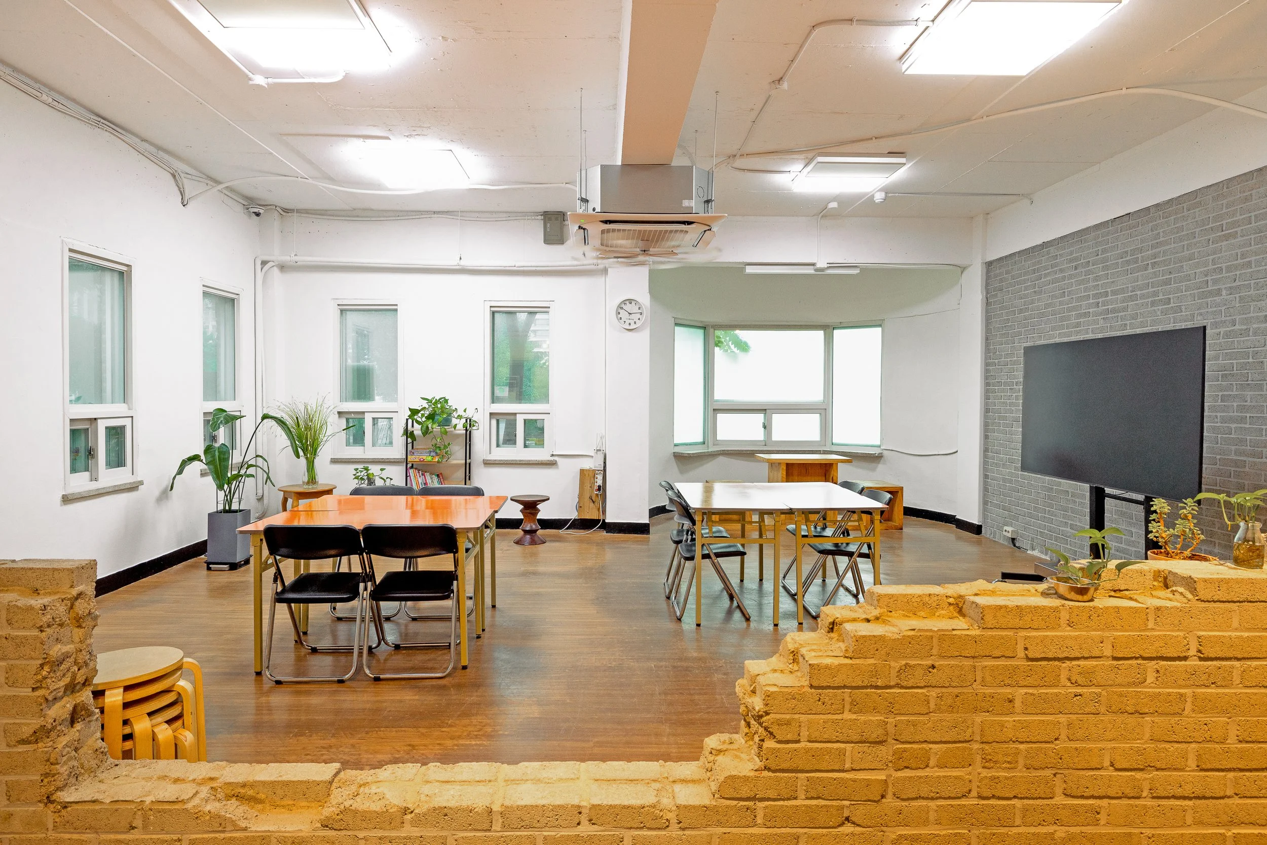 An interior space with multiple tables and chairs, potted plants, a large television on a brick accent wall, and several windows allowing natural light.