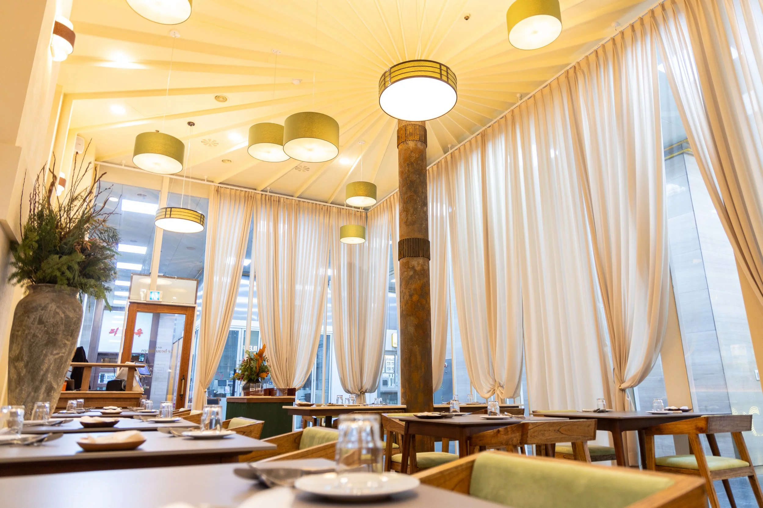 Interior of a restaurant with wooden tables, green chairs, large beige curtains, hanging ceiling lights, and a tall decorative column.