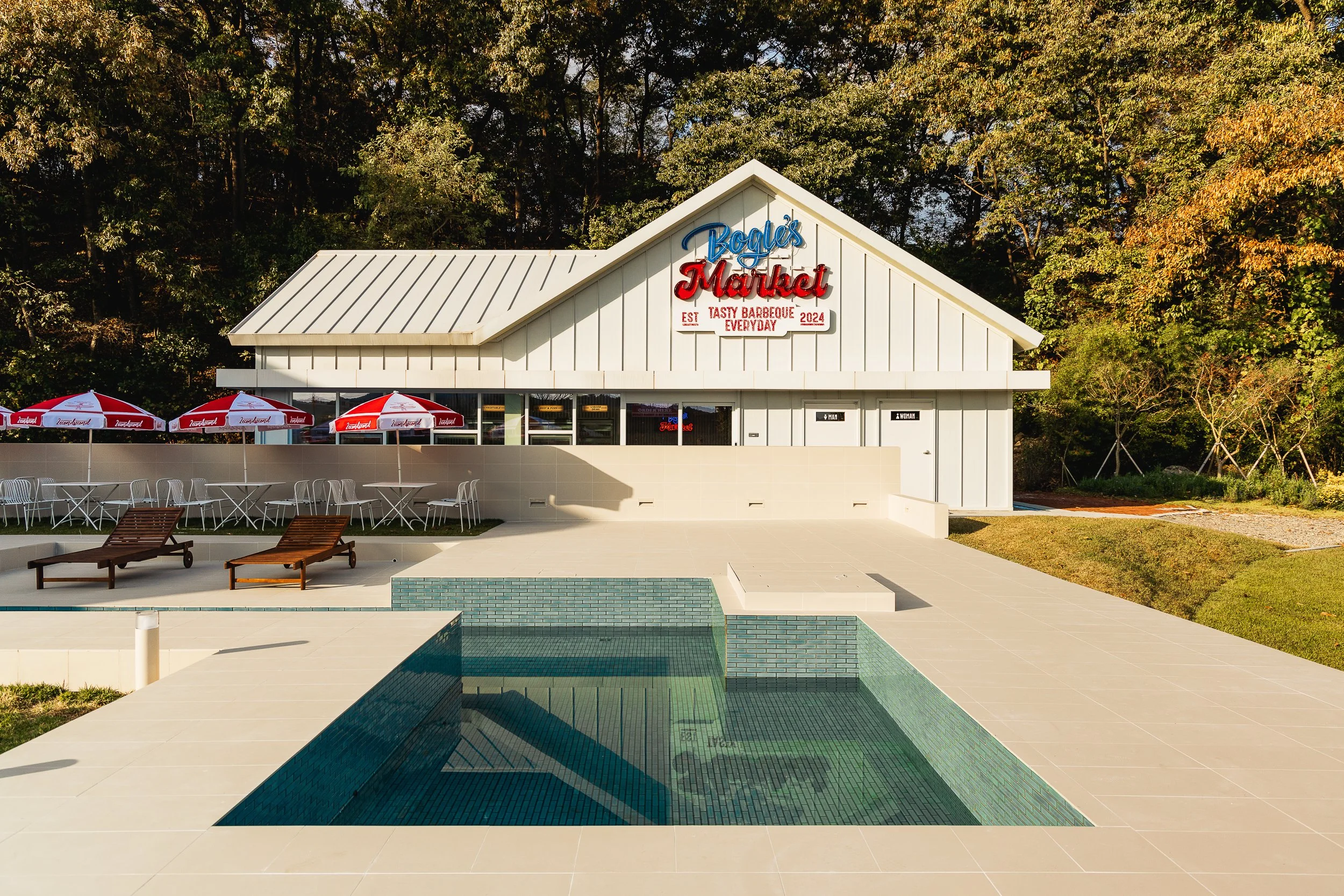 A poolside view of a white restaurant building with a sign that reads 'Buddy's Market' and features umbrellas, tables, and lounge chairs, surrounded by trees.