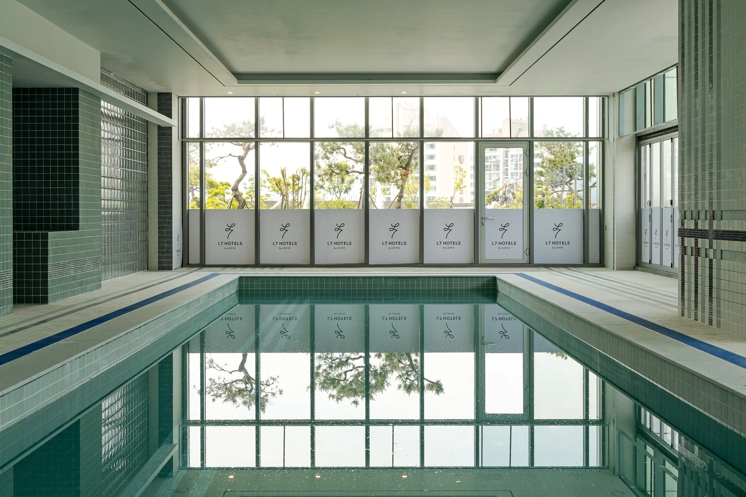 Indoor swimming pool with large windows overlooking trees and buildings outside, decorated with green and white tiles, with the logo 'L7 Hotels by Lotte' on the windows.