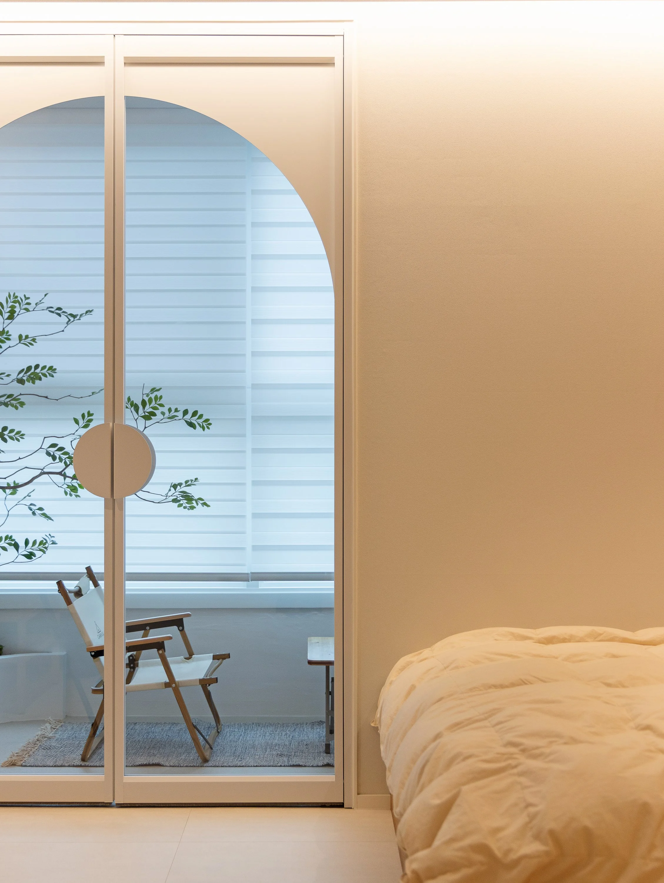 Bedroom with a bed covered in a beige comforter on the right, and a closet with sliding glass doors on the left, revealing a sitting area with a chair, a small table, a plant, and large windows with white blinds.