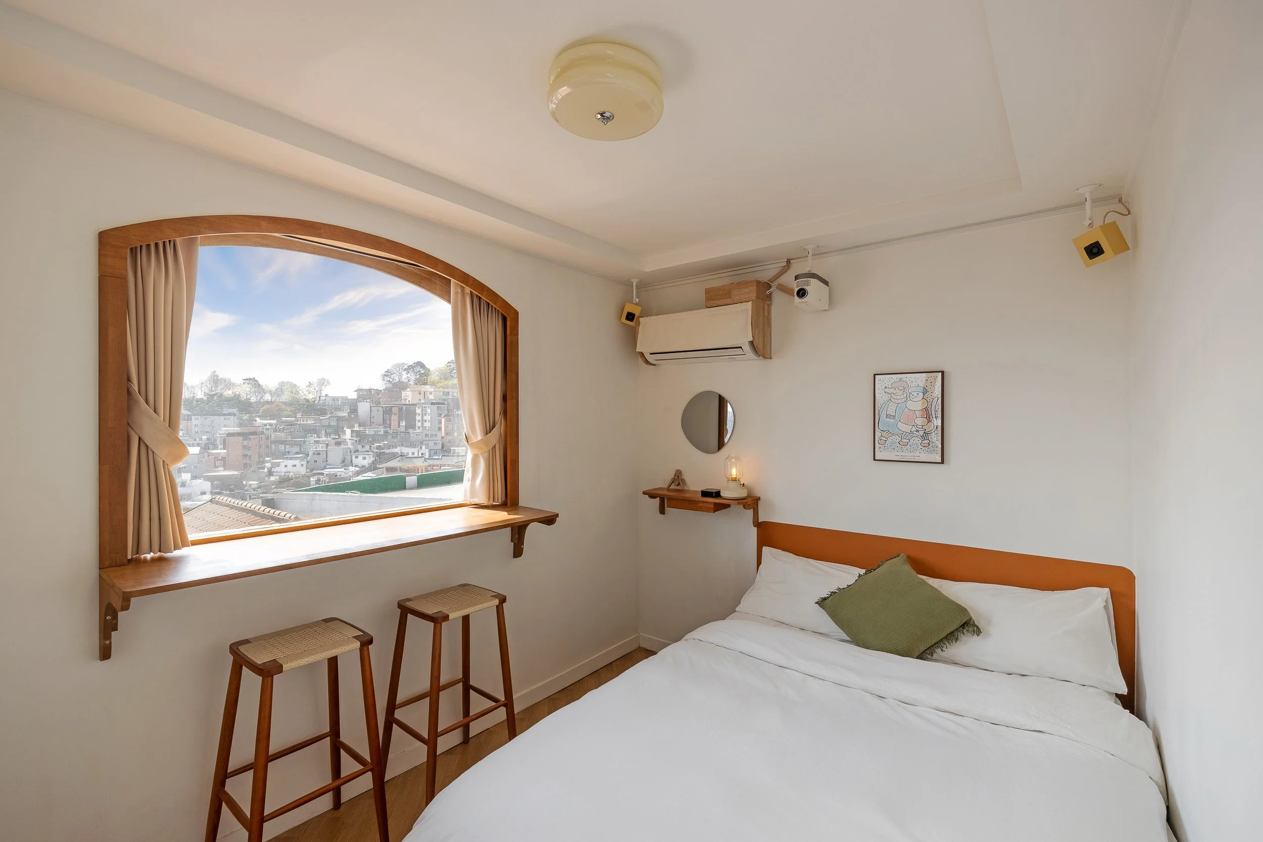 Cozy bedroom with a large window showing an urban view, a bed with white sheets and a green pillow, a small wall shelf with a lamp and decorative items, and a framed artwork on the wall.