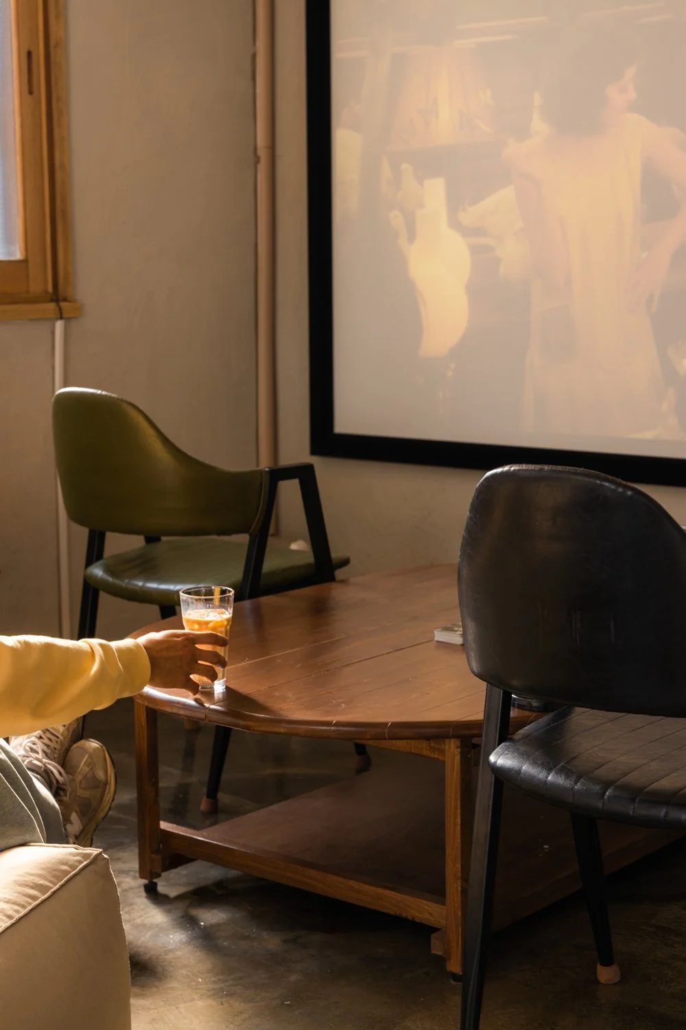 Person sitting on a couch holding a glass of orange drink, in a room with a brown wooden table, black and green chairs, a window, and a large projector screen showing a scene from a movie or TV show.