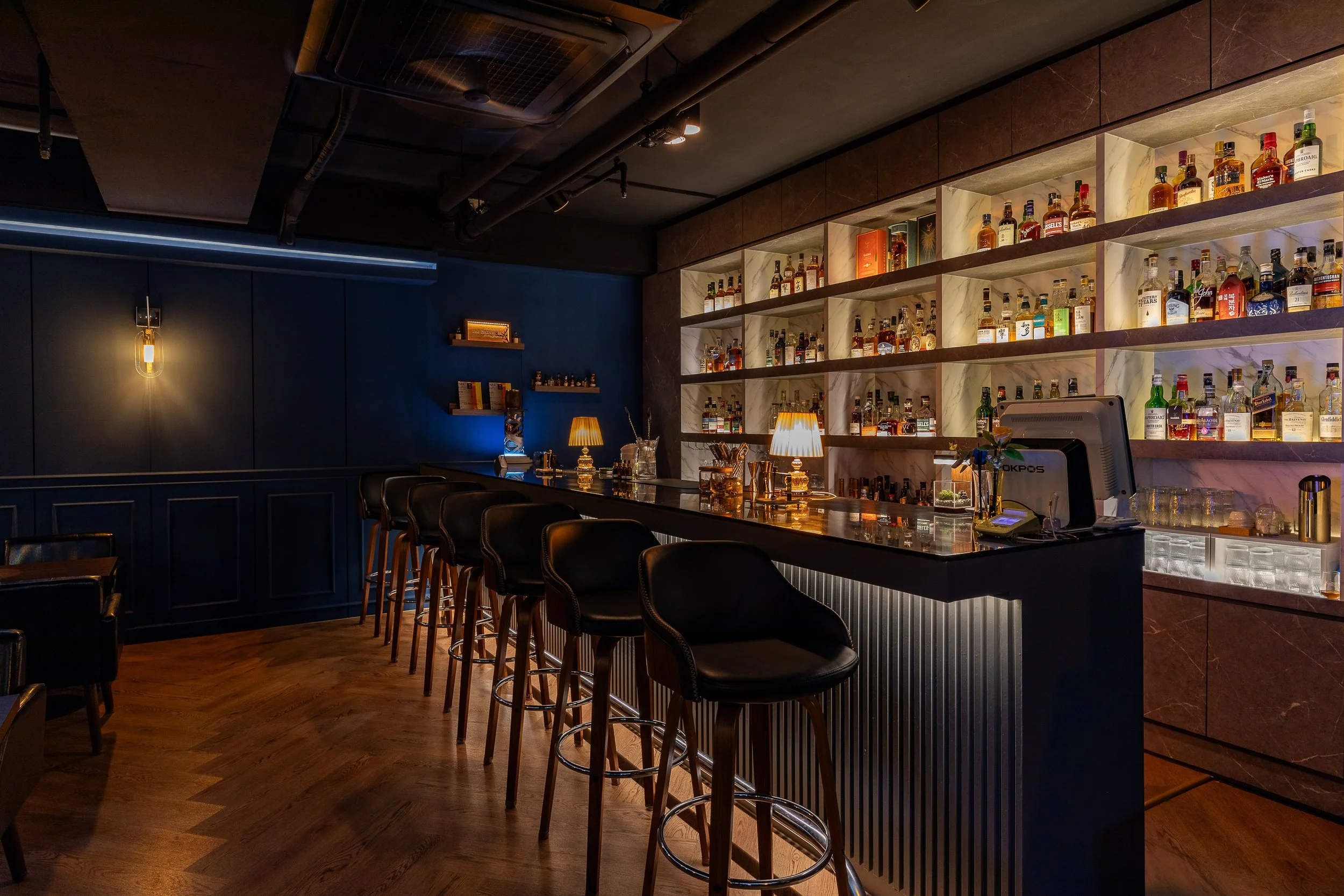 Interior of a dimly lit bar with a row of black barstools at the counter, shelves filled with liquor bottles, and warm lighting.