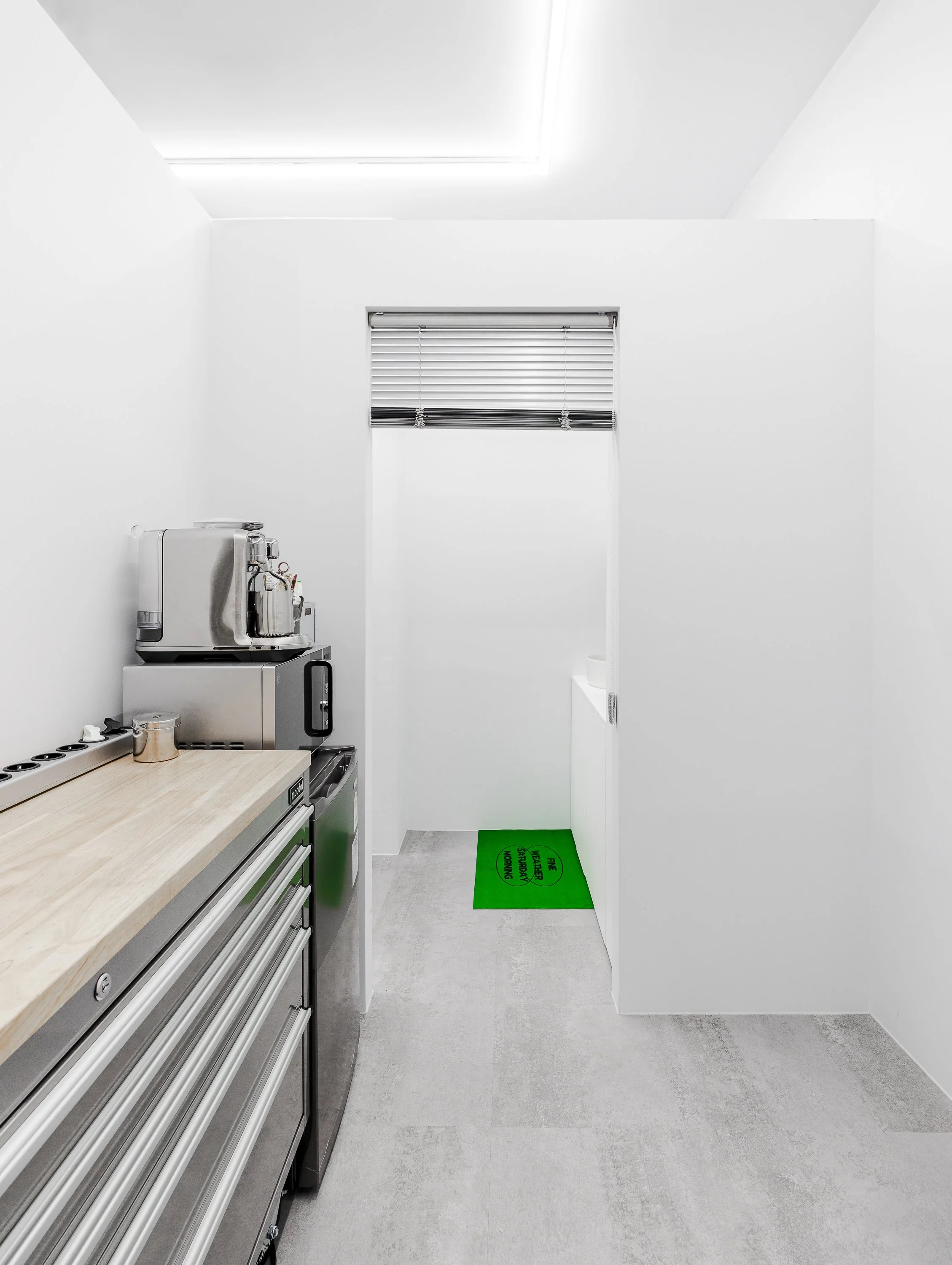 A modern, minimalist kitchen with a light wood countertop, a stainless steel coffee machine, a microwave, a trash bin, a window with blinds, and a small green mat on the floor that says "Please wash your hands".