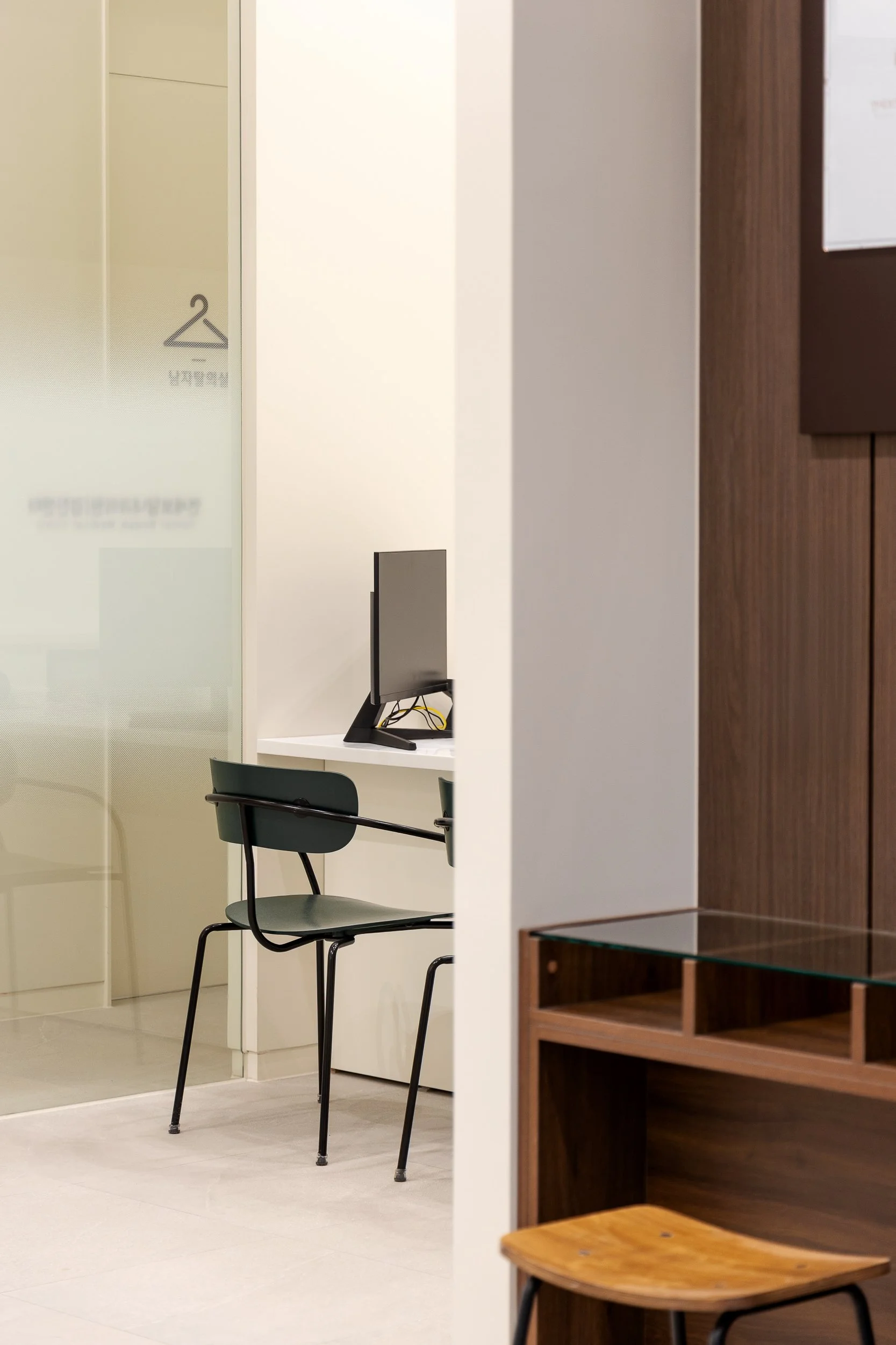 A small office area with a black chair, a computer monitor on a white desk, and a wooden cabinet with glass shelves in a modern, minimalist room.