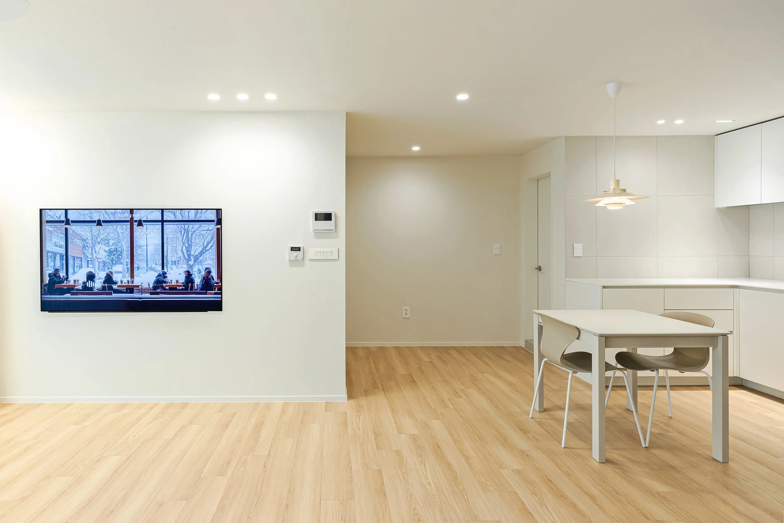 Minimalist living space with light wood flooring, white walls, a wall-mounted television displaying people at a snowy outdoor cafe, and a small dining table with chairs near the white kitchen cabinets.