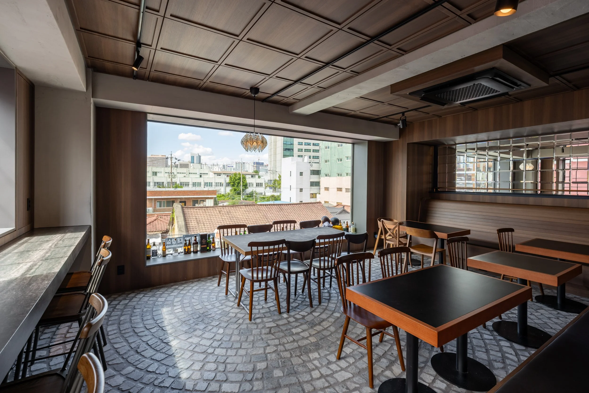 Interior of a modern restaurant with black tables, wooden chairs, and a large window with a city view. The ceiling has a wooden panel design with track lighting, and some bottles are placed on the window sill.