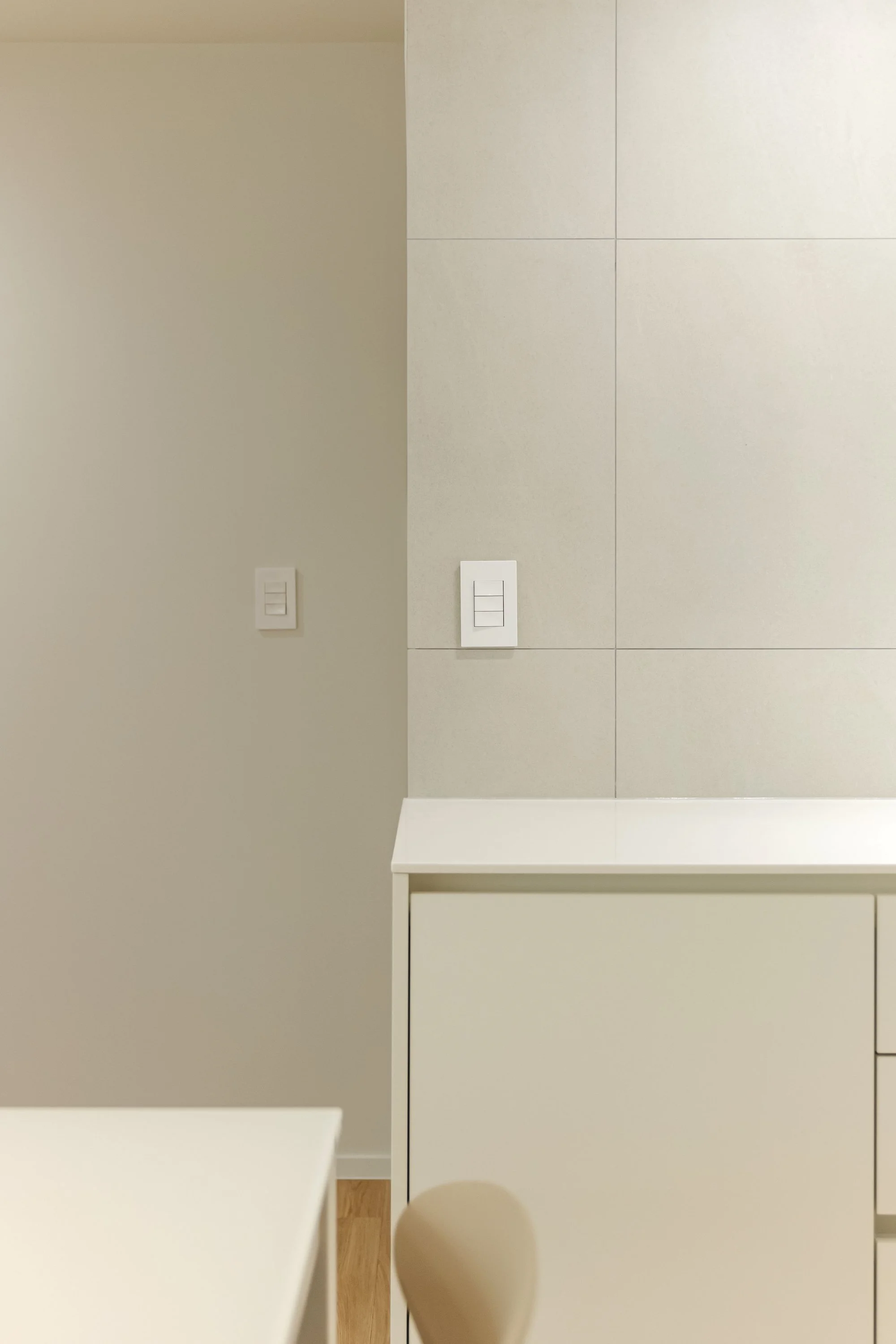Part of a modern kitchen with a beige wall, white electrical switches, and a white cabinet. A chair and a tiled wall are also visible.
