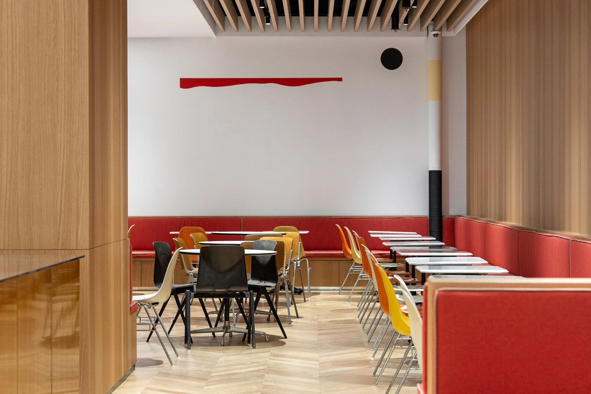 Empty restaurant with various chairs and tables, red banquettes along the walls, light wood walls, and ceiling with exposed beams.