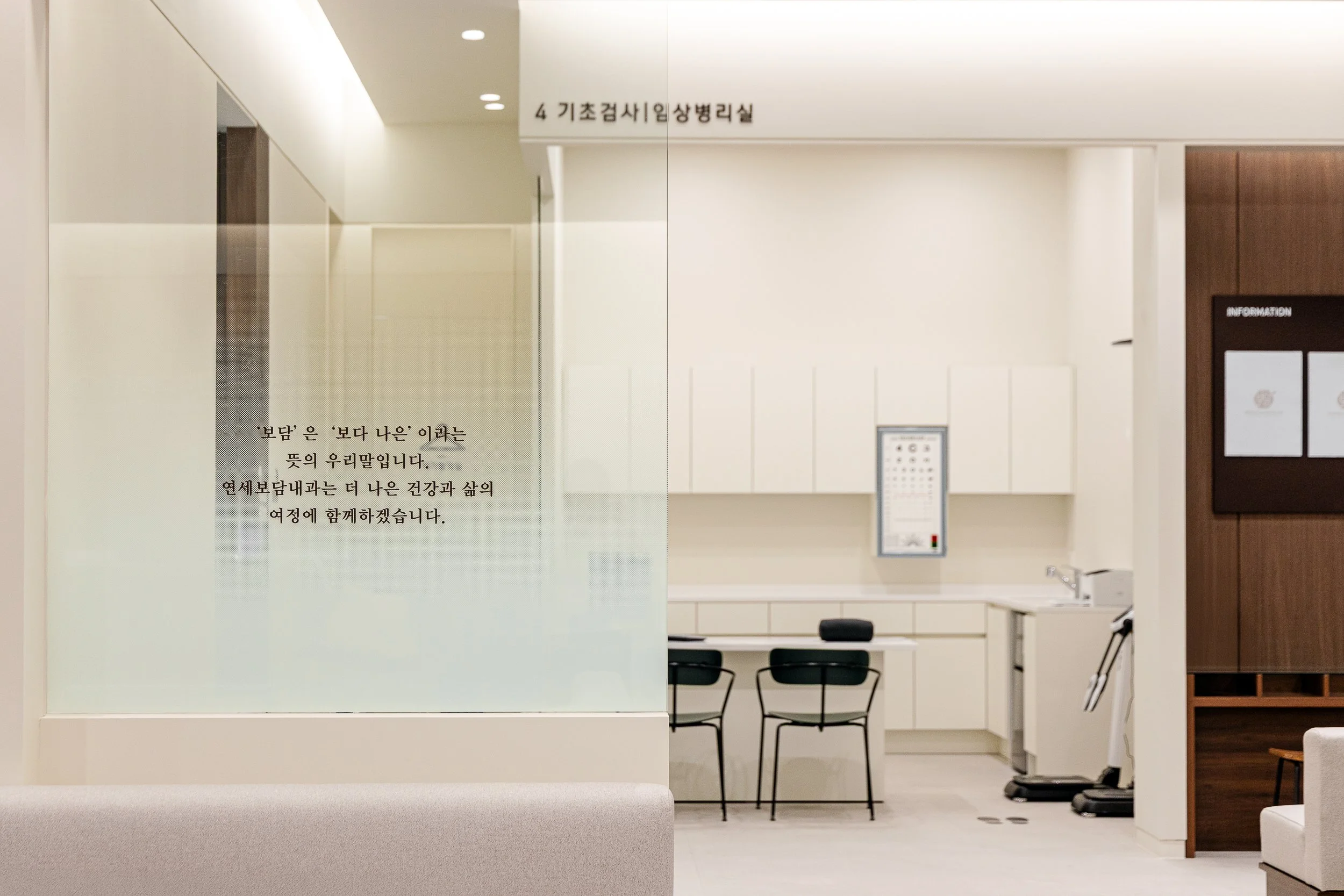 Modern medical examination room with white walls, black chairs, and a frosted glass partition with Korean writing, featuring a white cabinet, a keyboard, and medical equipment.