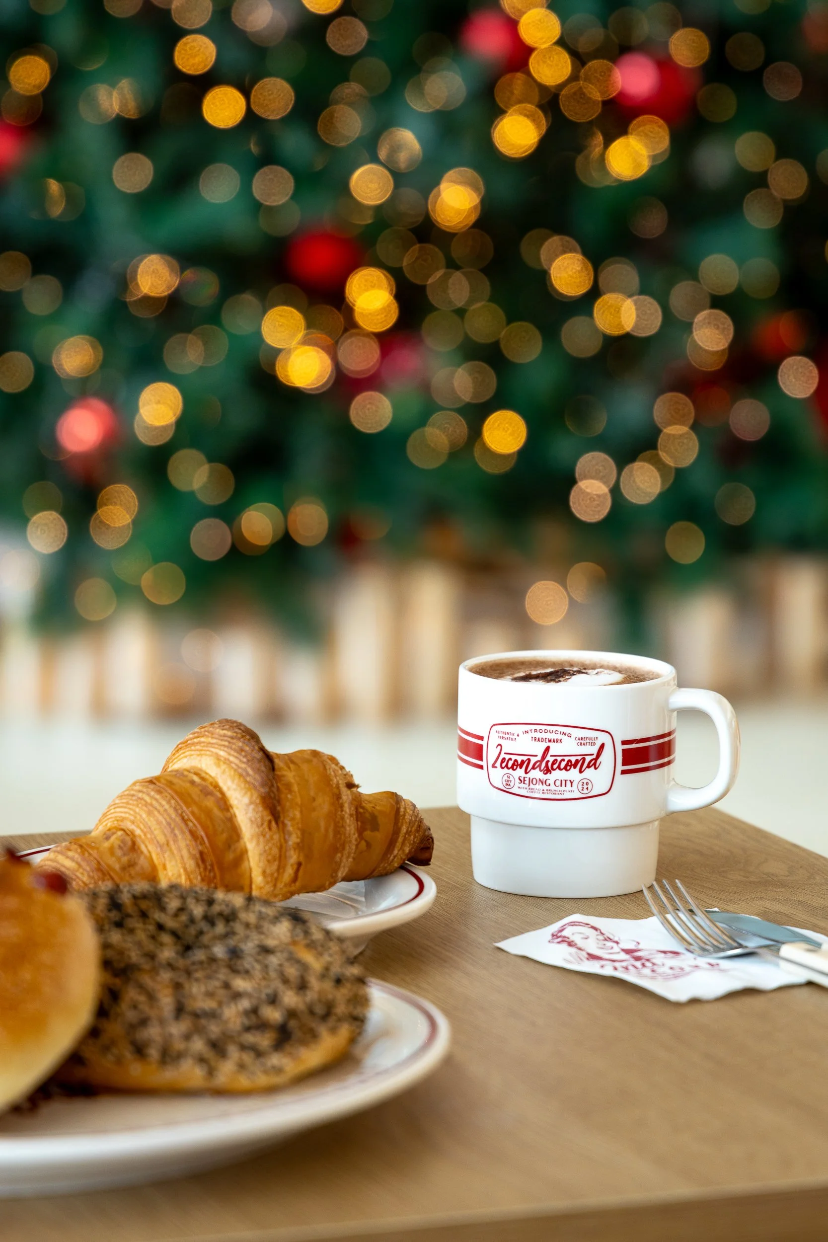 A breakfast scene with a croissant and a pastry on plates, a mug of hot chocolate or coffee, and a Christmas tree with blurred lights in the background.