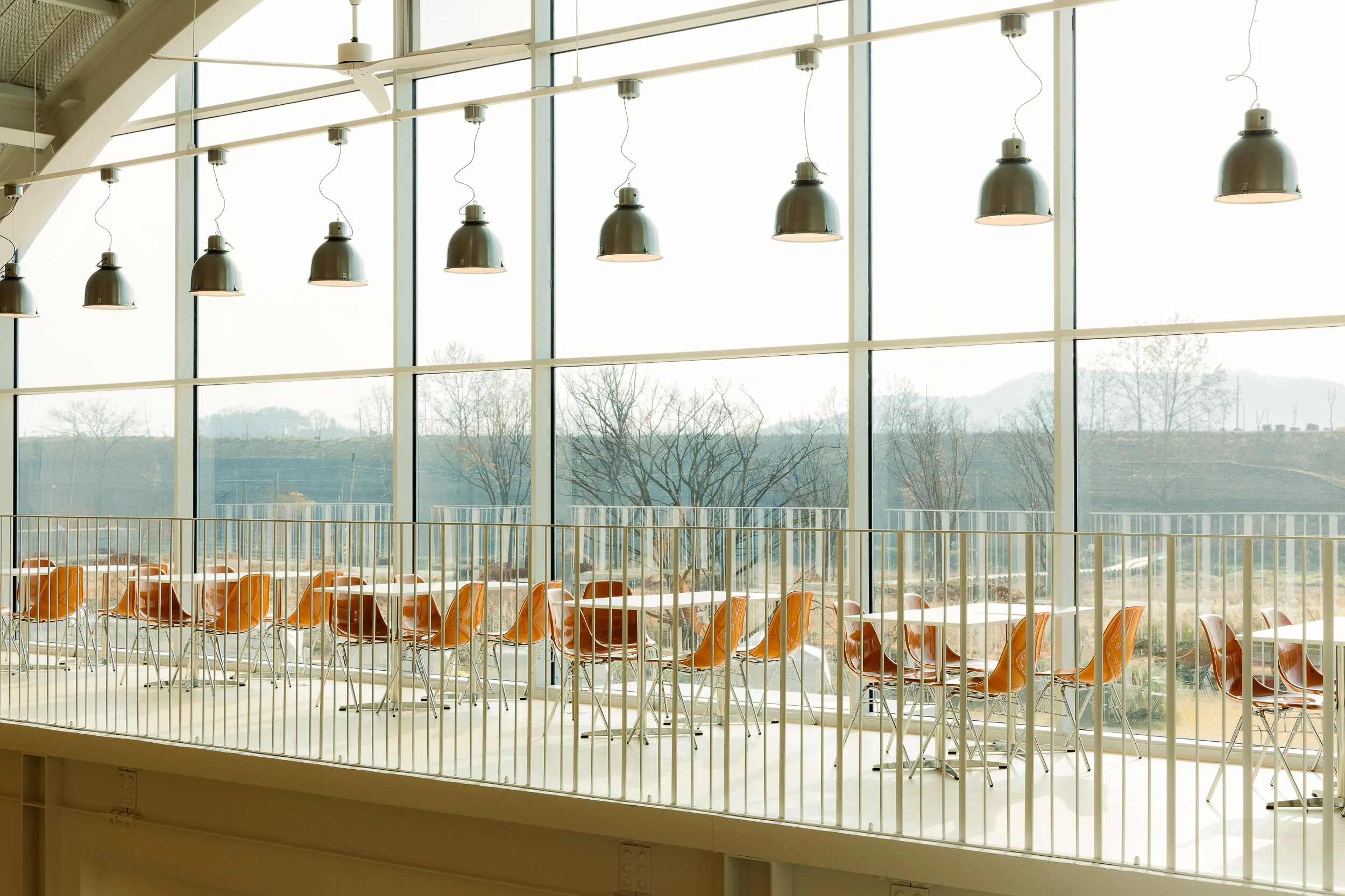 Empty indoor space with large glass windows, orange chairs, white tables, and hanging pendant lights.
