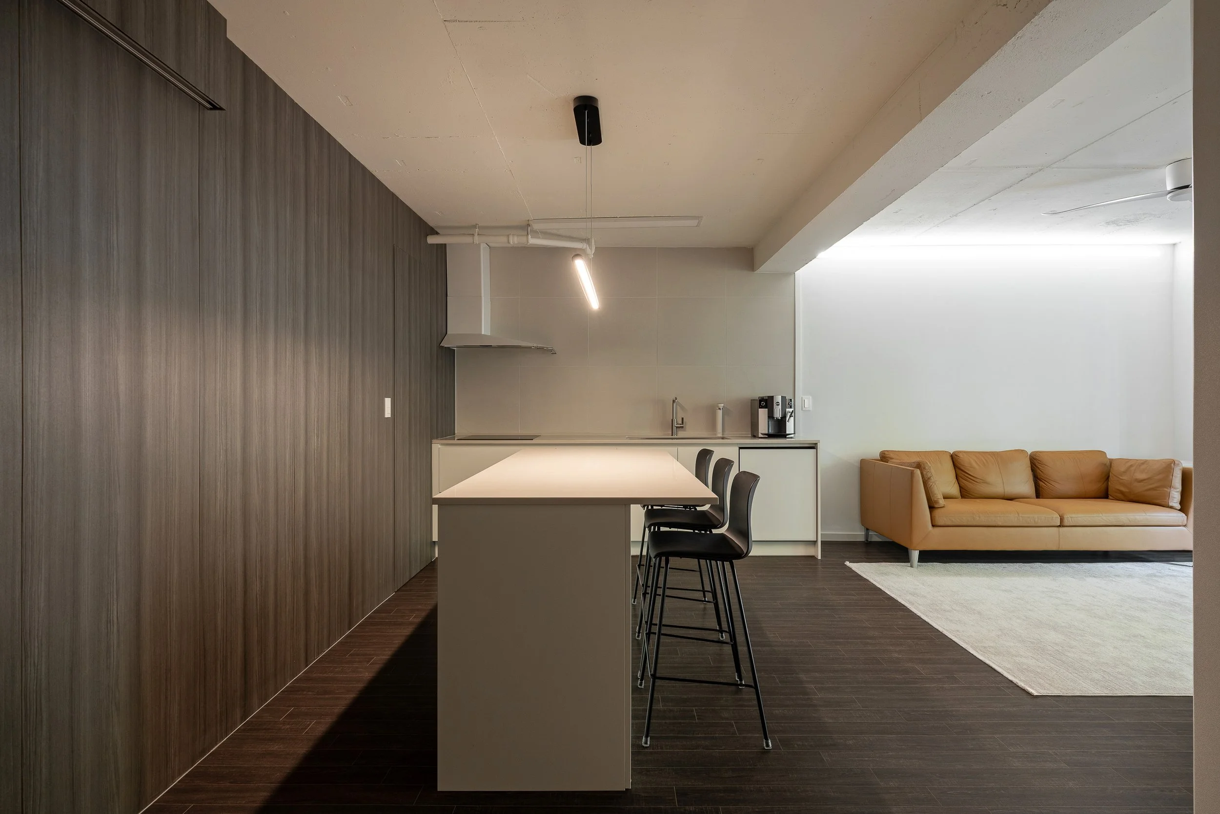 Modern kitchen and living area with dark wood flooring, a white countertop island with four black barstools, beige couch, and a white area rug in a minimalist design.
