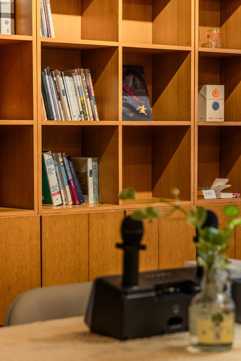 Empty wooden bookshelf with scattered books, a box, and decorative items, with a blurred plant and a black speaker on a table in the foreground.