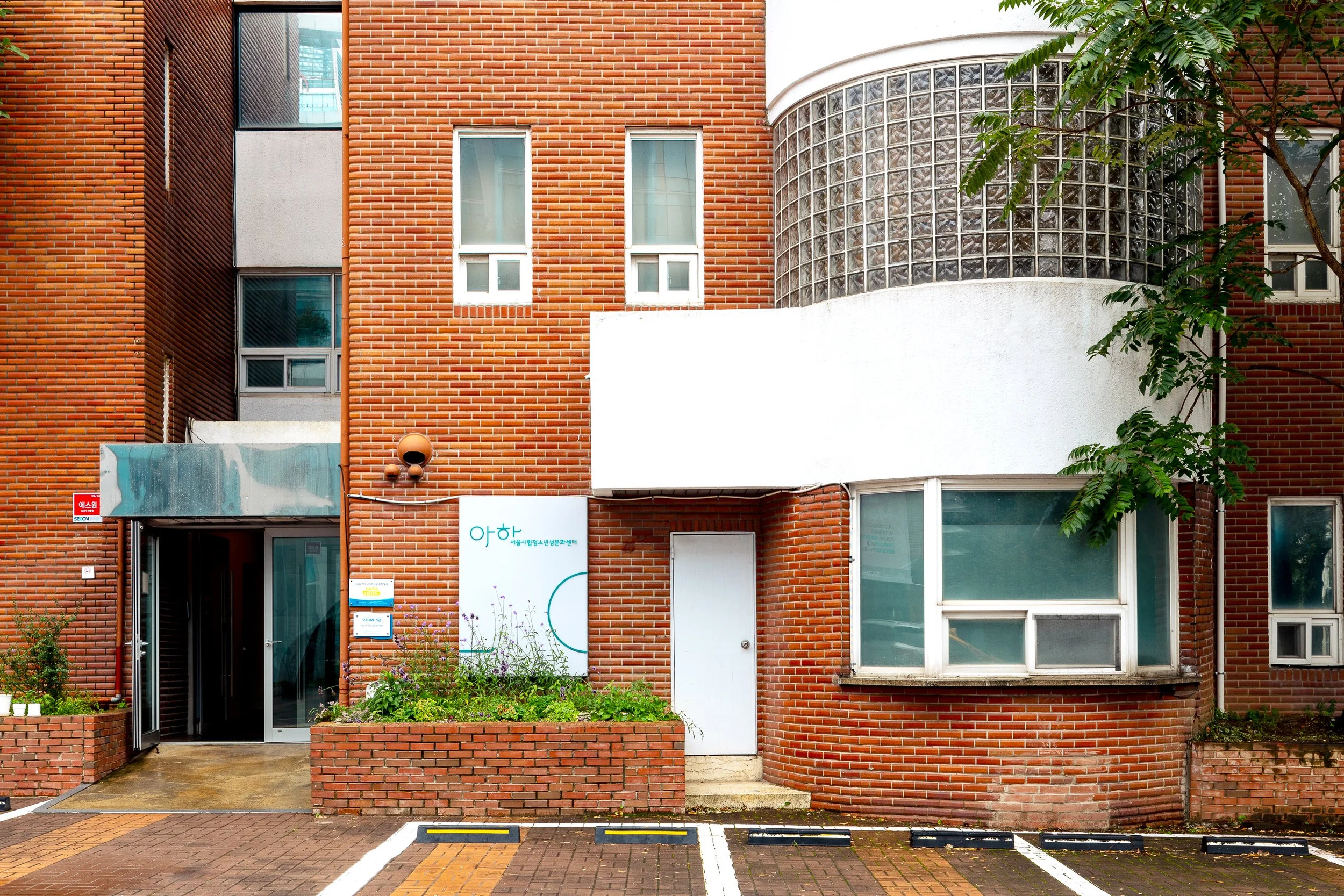 Red brick building with a white door, a parking space with yellow markers, and a garden with plants. There are windows and exterior walls made of red bricks, and a curved glass block section protruding from the building.