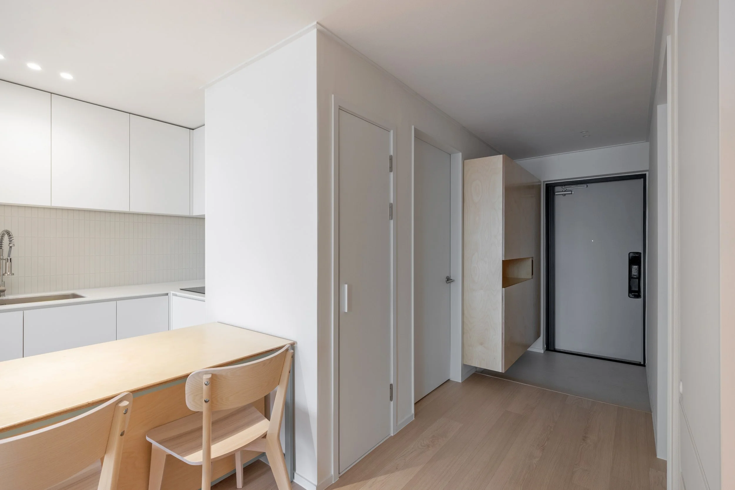 Interior view of a modern apartment with a white kitchen, wooden dining table and chairs, and an entryway with a black door.