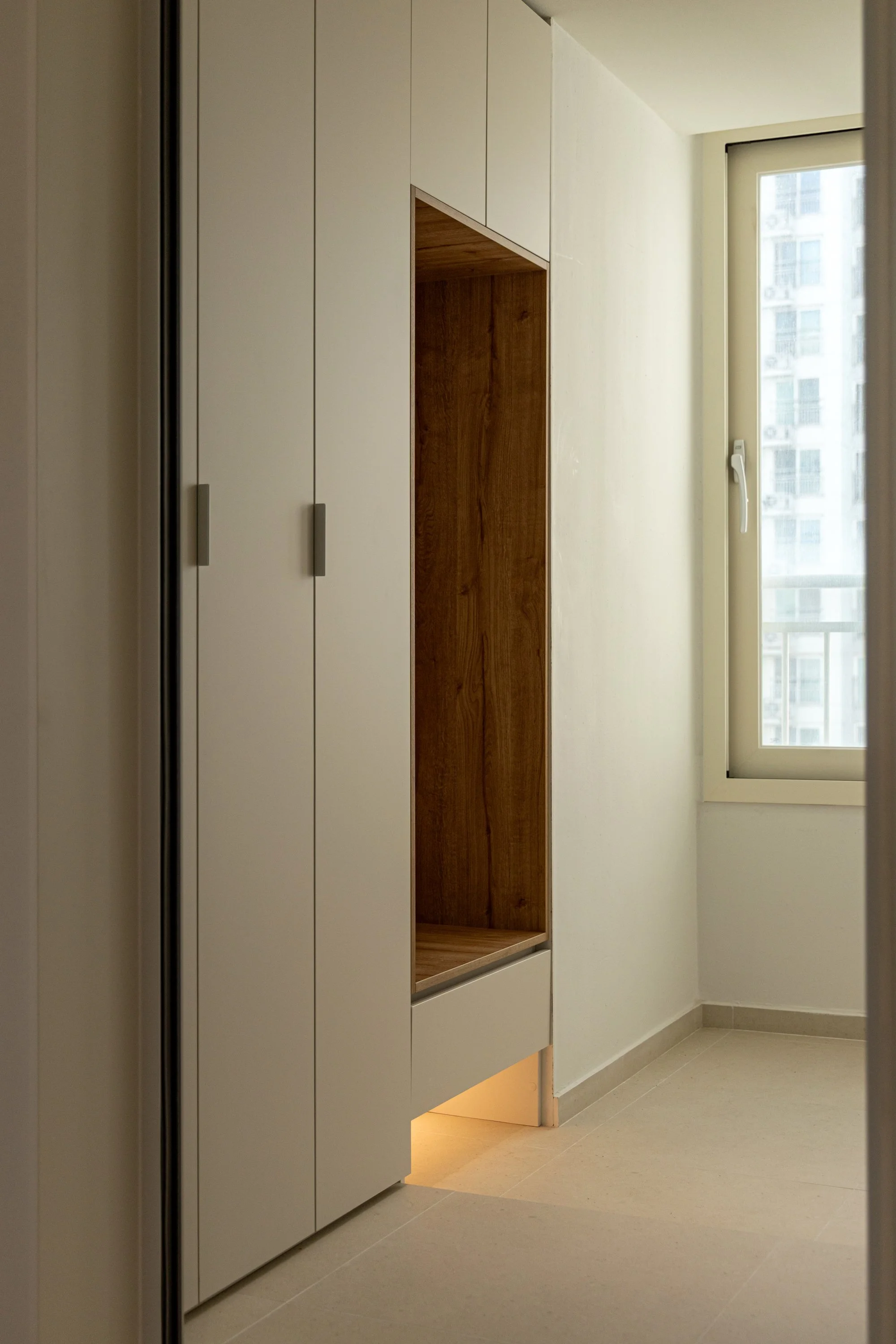 Modern interior space with built-in white closet cabinets, wooden open storage niche, and a large window letting in natural light.