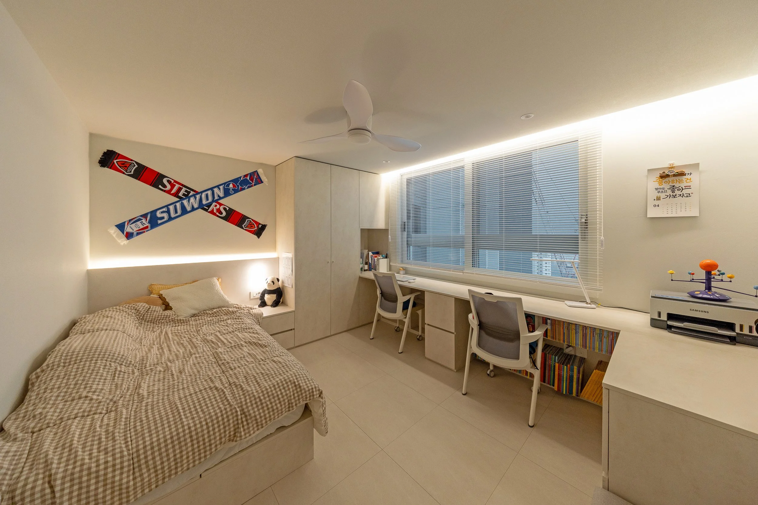 A bedroom with a bed on the left side, decorated with a panda plush and framed sports scarves above. The right side features a long white desk with two chairs, a window with blinds, a bookshelf underneath, and a corner with a printer and a test tube 