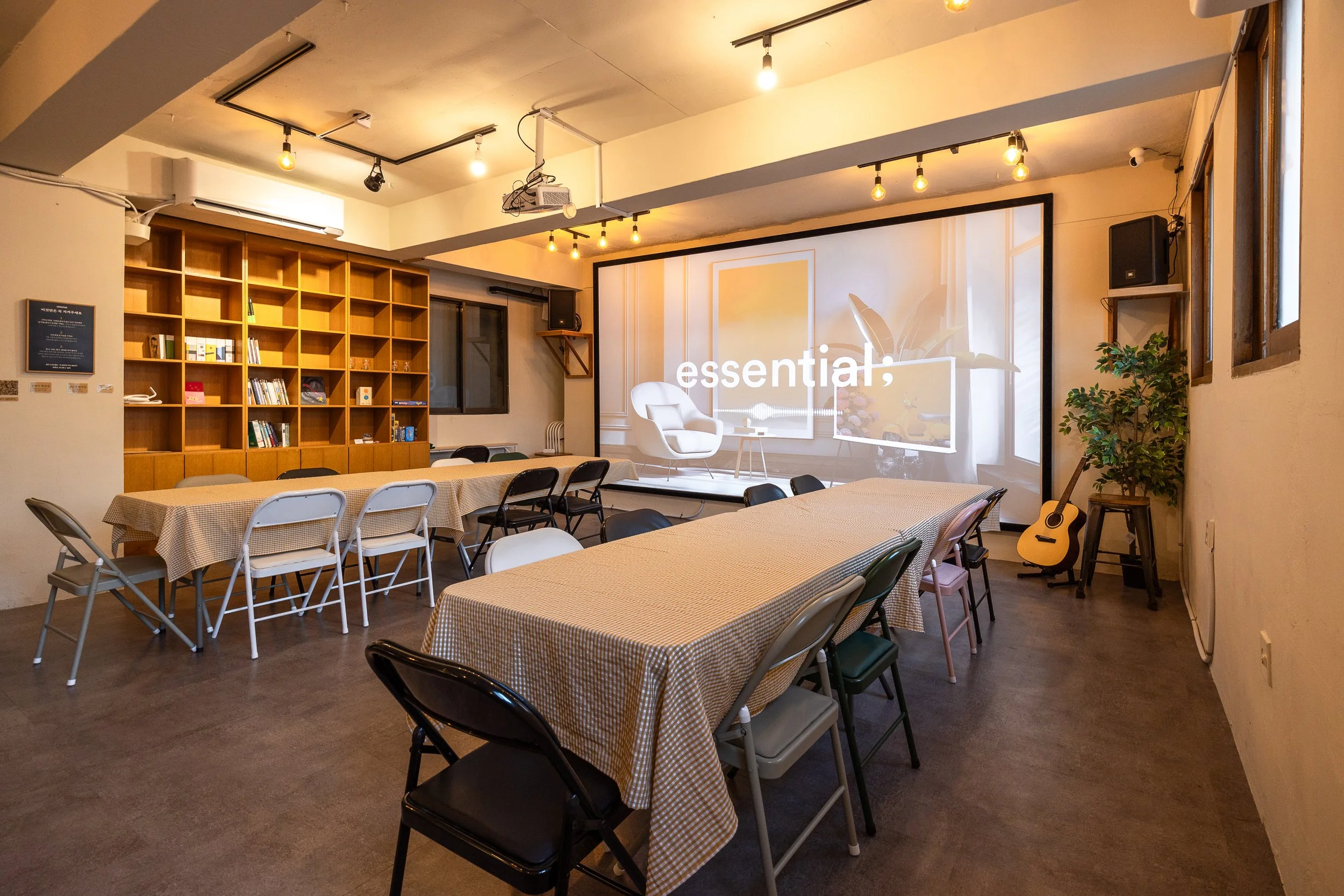 A cozy, well-lit room with tables covered in yellow checkered tablecloths, surrounded by various chairs, a large bookshelf filled with books and decor, a projector mounted on the ceiling projecting a presentation titled 'essential:', a sitting area on the stage with a white chair, a guitar leaning against a plant on a stool, and windows bringing in natural light.