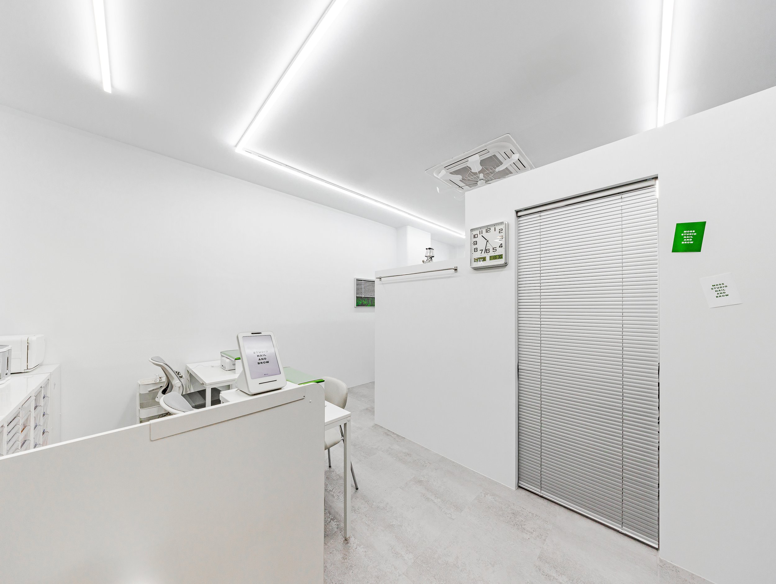 A minimalistic, white hospital or clinic room with a computer on a desk, a chair, a wall clock, and window blinds.
