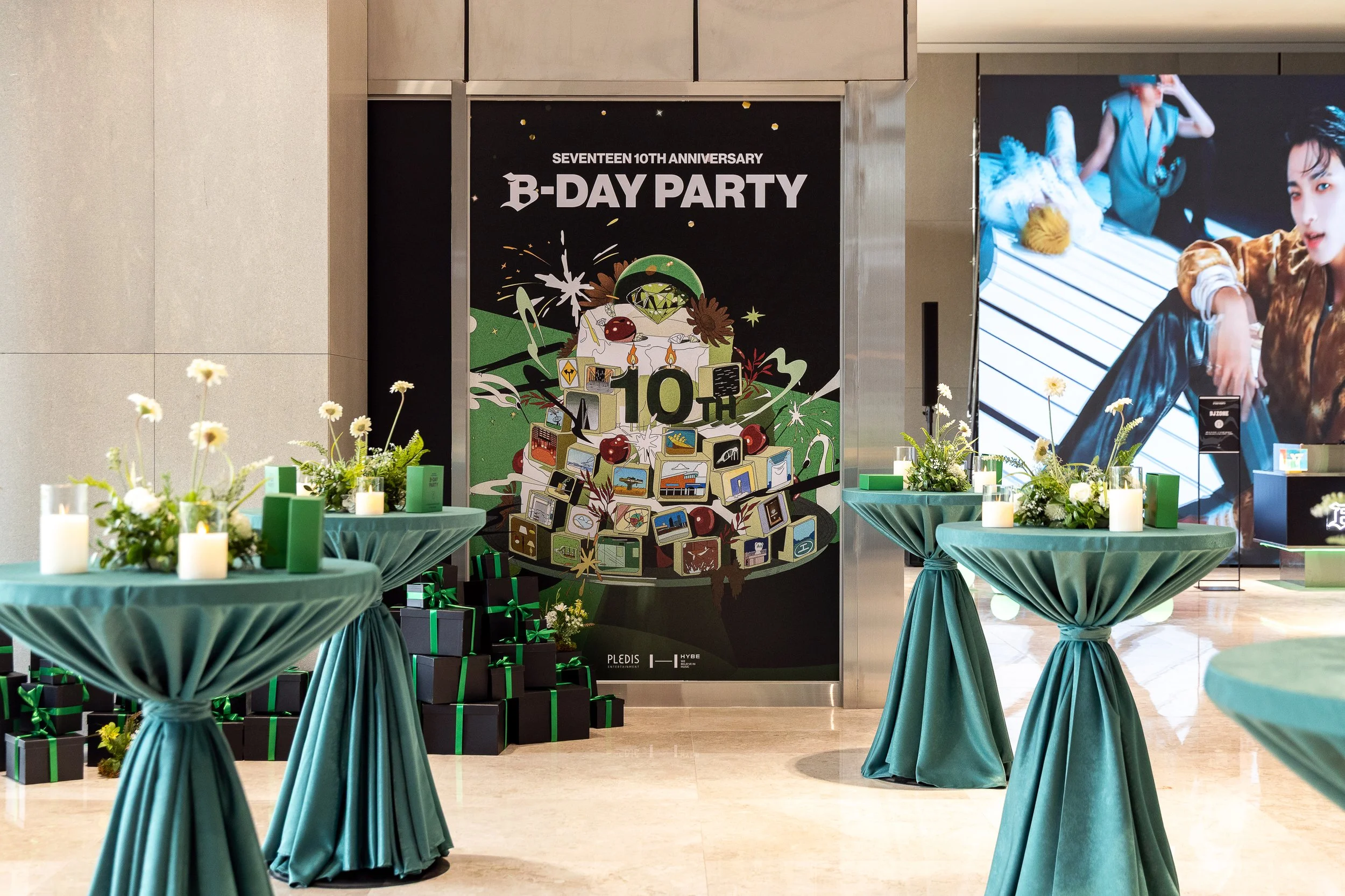 Decorated tables with flower arrangements and candles at a 10th anniversary birthday party event in a mall or lobby area, with a large poster in the background and a digital display.