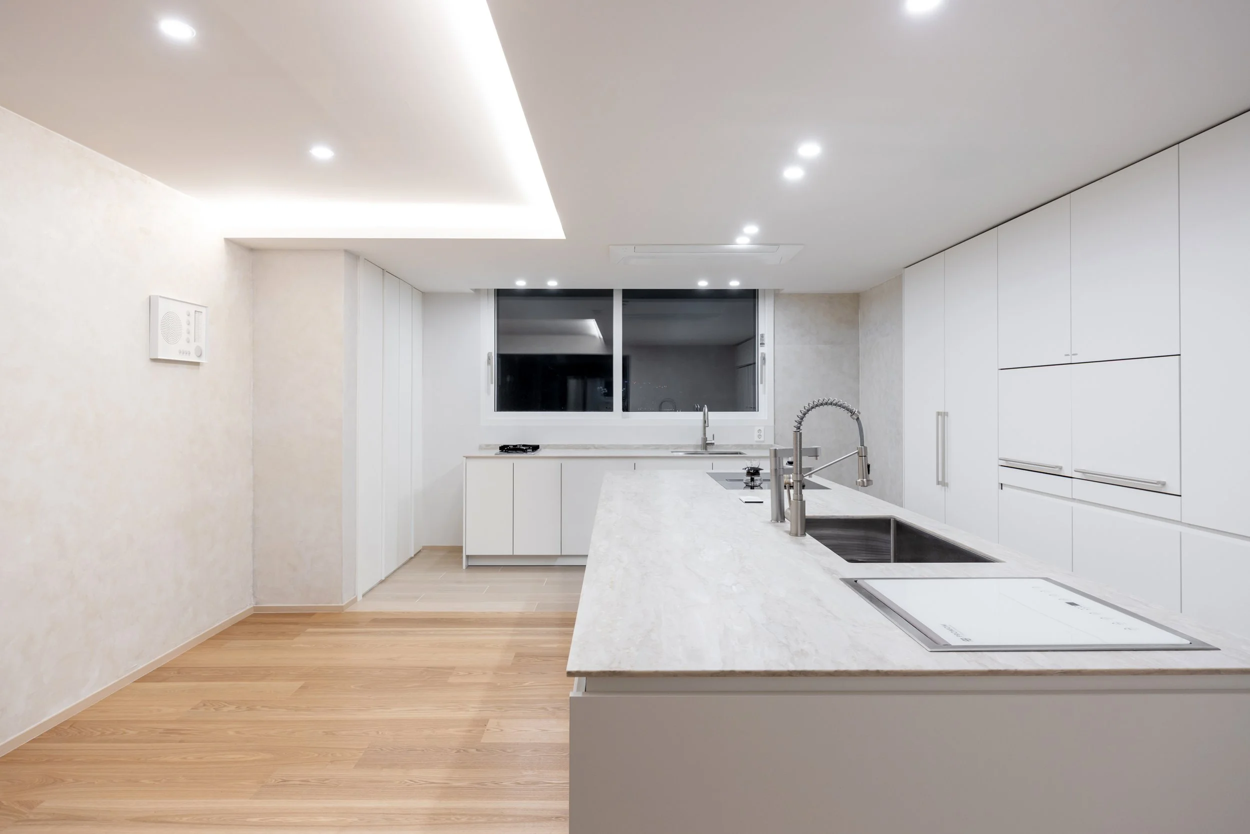 Modern white kitchen with marble countertops, sinks, and stainless steel faucets, illuminated by ceiling lights, with a window showing a dark night outside.