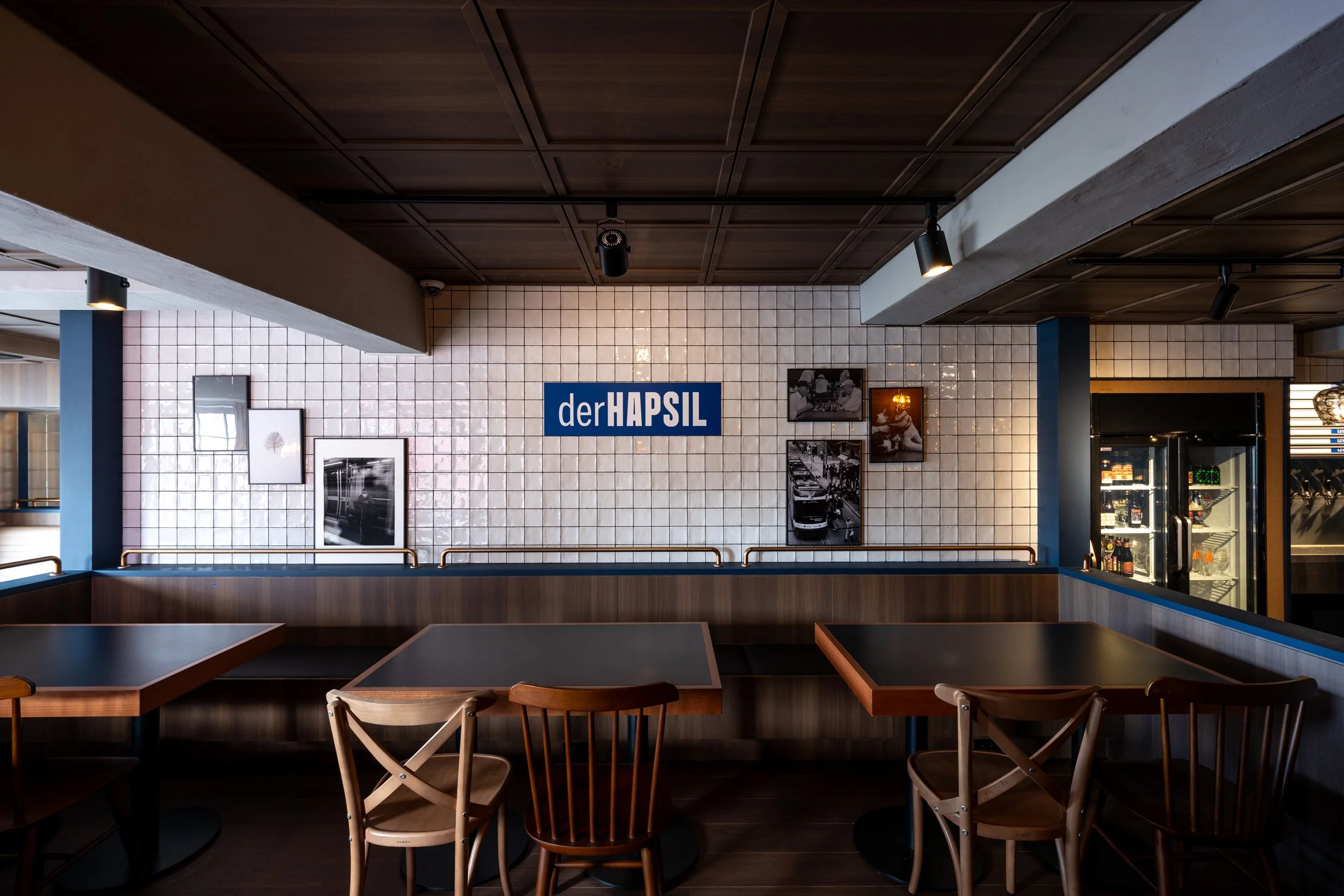 Interior of a restaurant or bar with wooden tables and chairs, white tiled wall with framed pictures, a sign that reads 'der HAPSIL', and a beverage refrigerator.