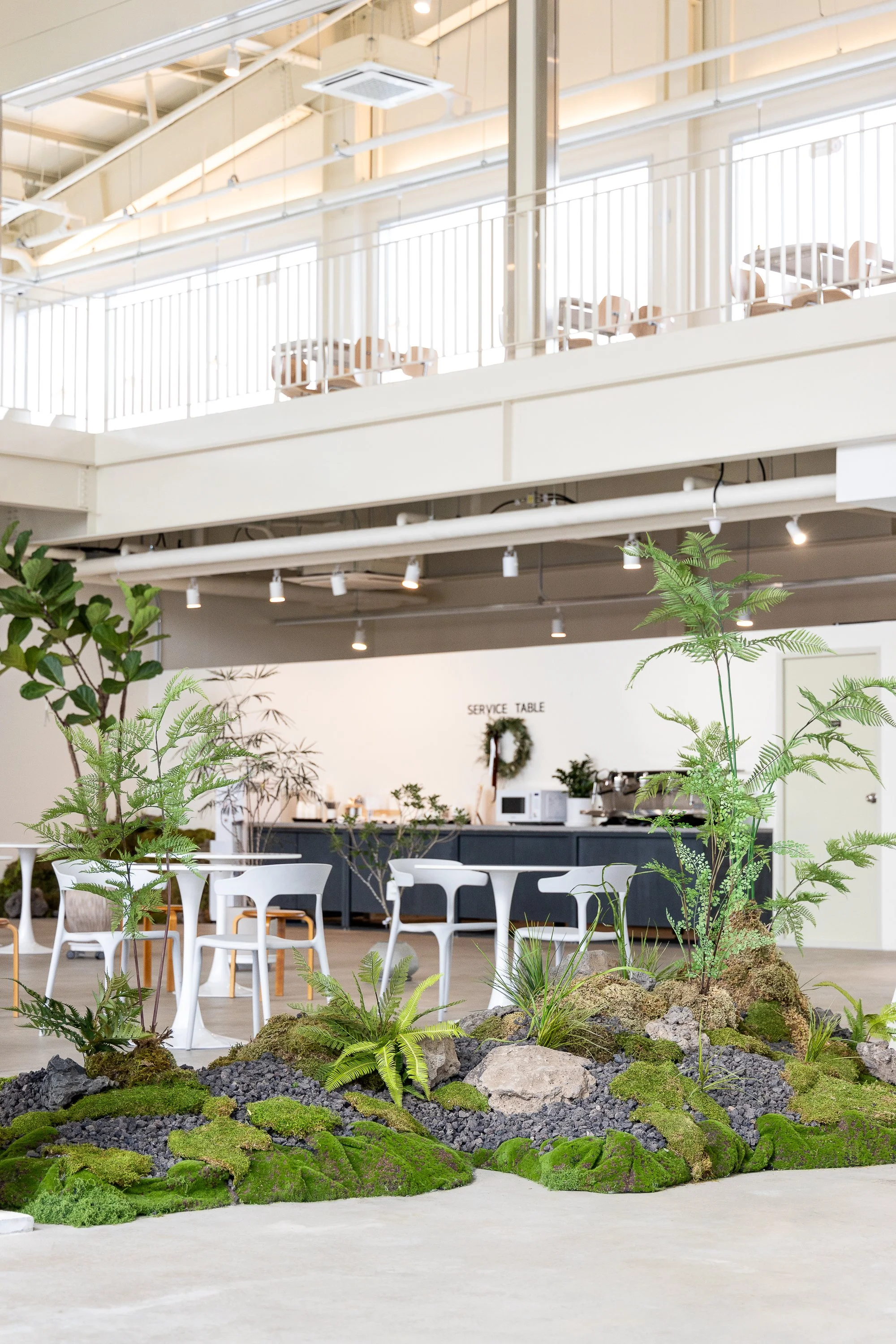 Indoor space with a moss and plant garden in the foreground, white chairs and tables in the middle, and a service area with a microwave, coffee machines, and a wreath on the back wall. The upper level has a balcony with chairs and tables. Modern and 