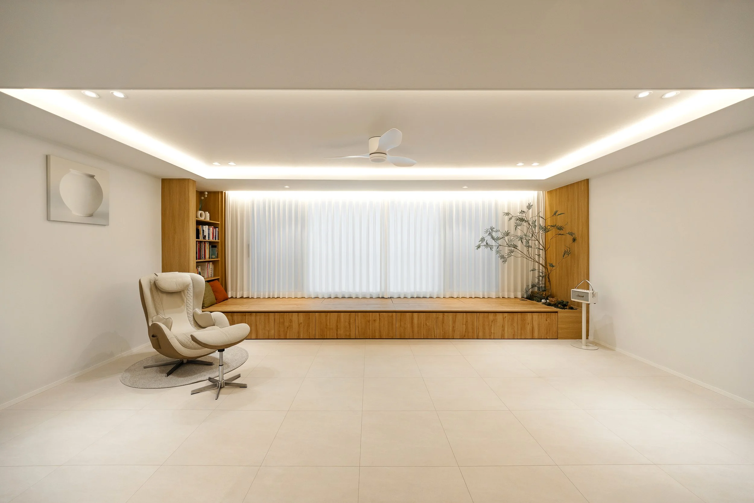 Minimalist living room with white walls, ceiling, and floor tiles, featuring a white armchair with a curved design on a small gray rug, a built-in wooden window seat with white curtains, a corner bookshelf filled with books, a decorative vase on the 