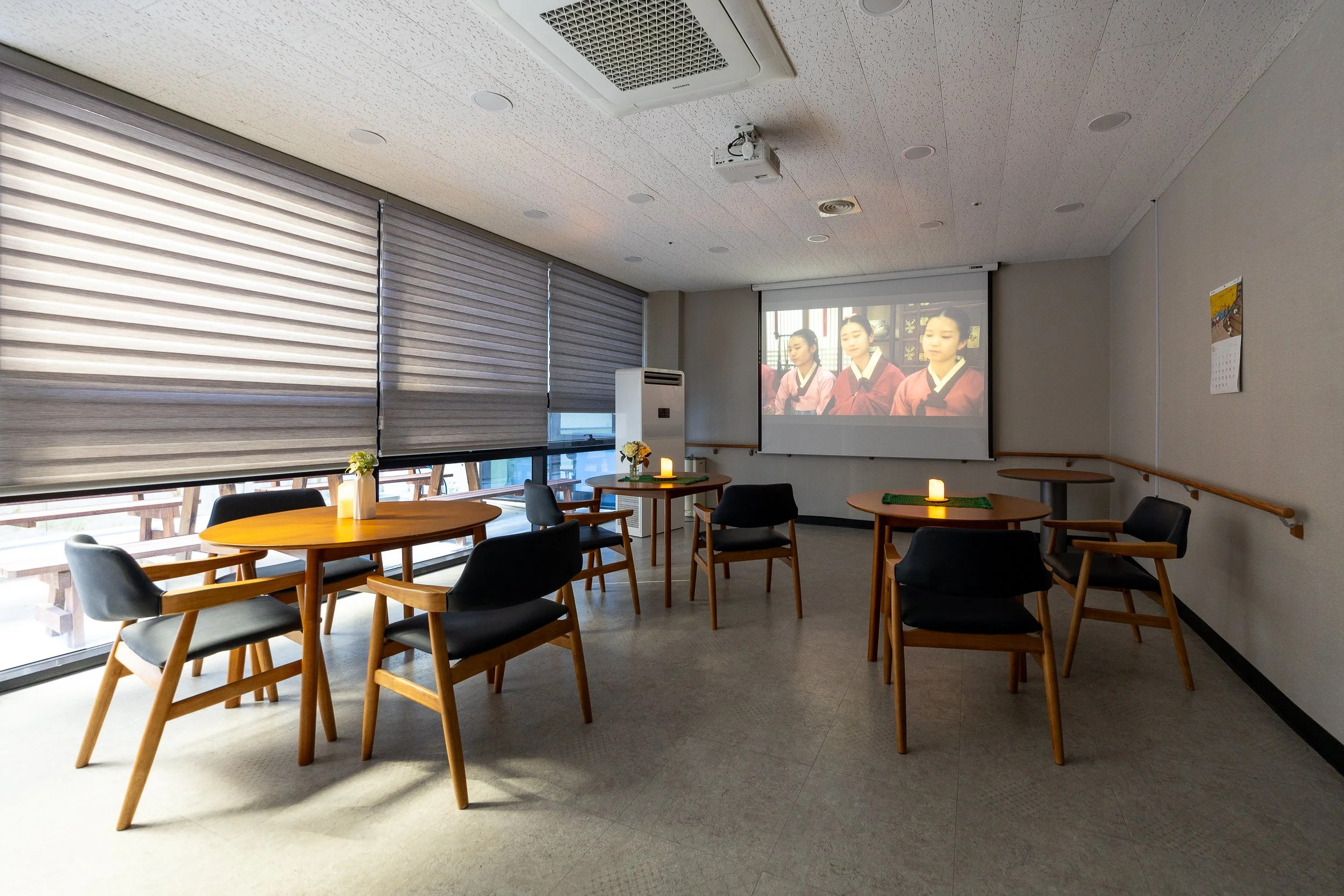 Empty room with five round wooden tables, each with a small candle, two of which have green table runners. Six black and wooden chairs with armrests are arranged around the tables. Large windows with closed blinds occupy the left wall. A large screen