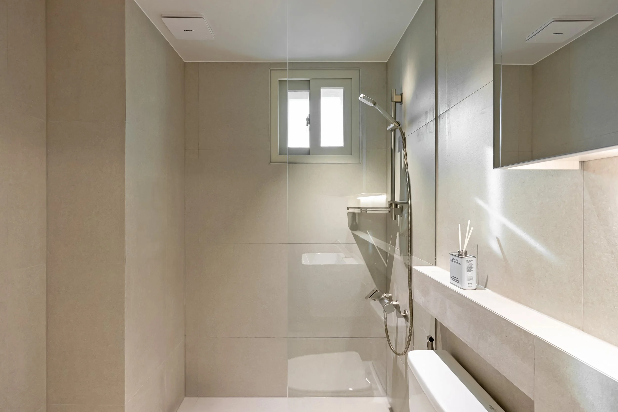 Modern minimalist bathroom with beige tiled walls, a small window, a sleek shower area with a handheld showerhead, and a white container with reed diffusers on a shelf.