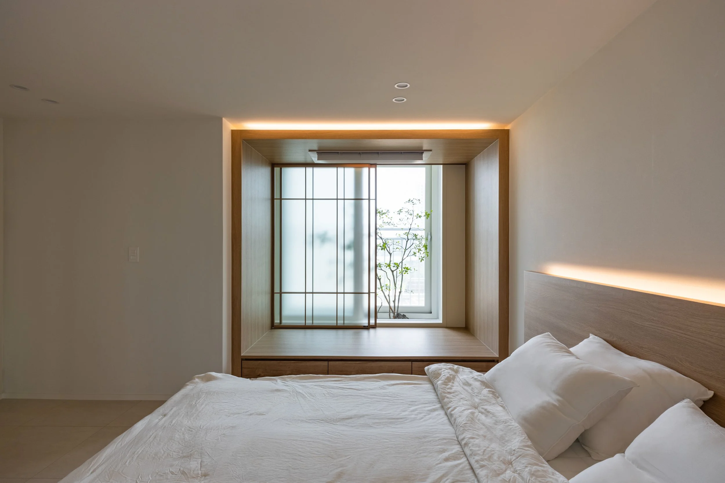 Modern bedroom with a wooden bed and white bedding, large window with frosted glass and sliding screen, a small potted plant outside, and minimal lighting.