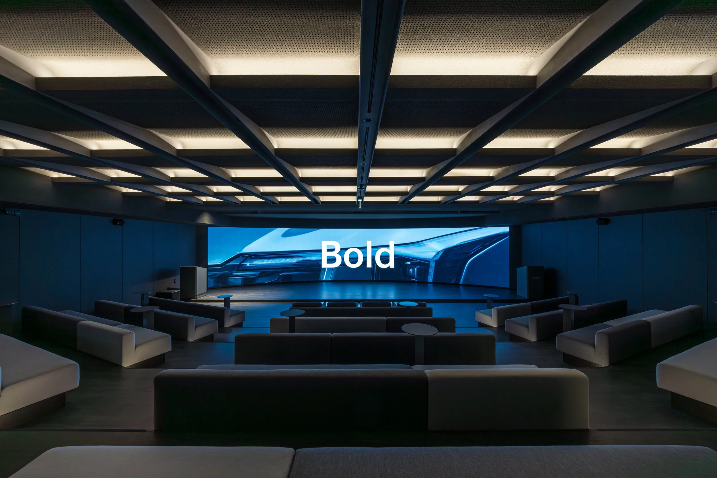 A modern presentation room with a large screen displaying the word 'Bold' over an image of a sleek car front. The room has a high ceiling with rectangular lighting panels and several white sofas and armchairs arranged facing the screen.