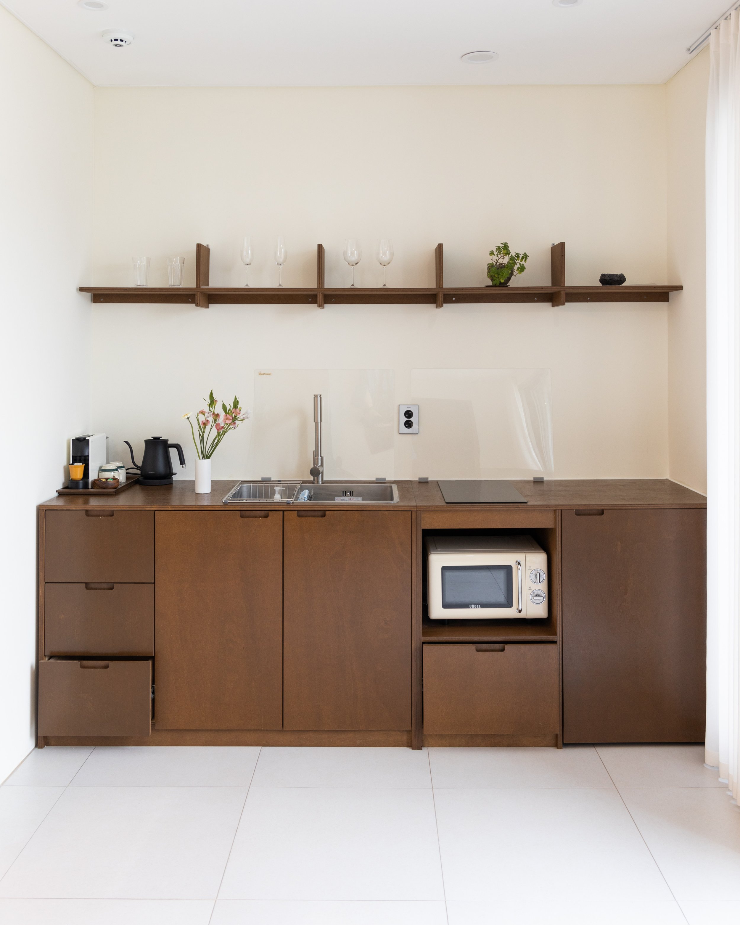 Minimalist kitchen with brown wooden cabinetry, a small microwave, a sink, a black teapot, and a white vase with pink flowers, with a wooden shelf holding glasses and plants on a light-colored wall.