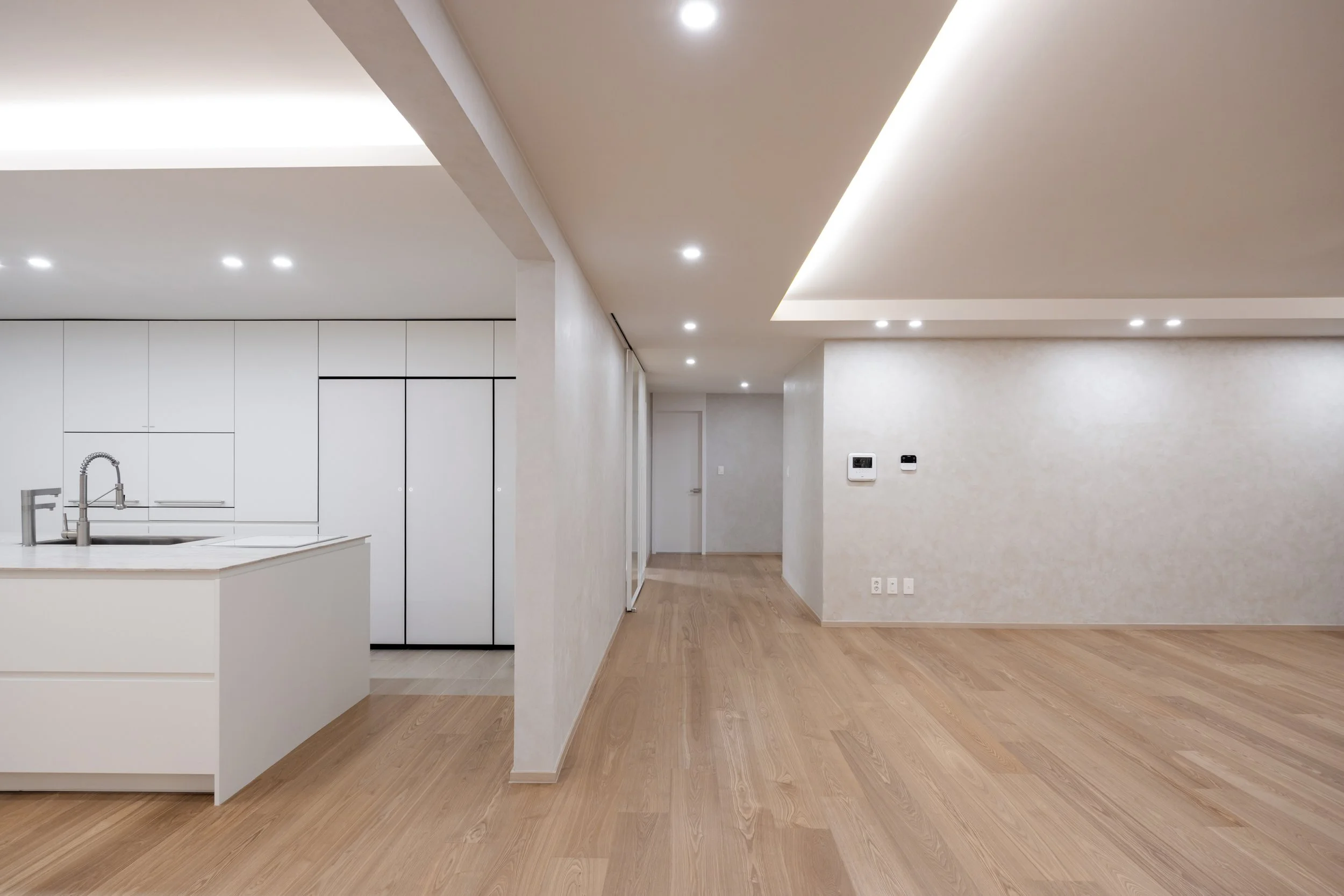 Empty modern living room with hardwood floors, white walls, recessed ceiling lights, and a white kitchen island in the background.