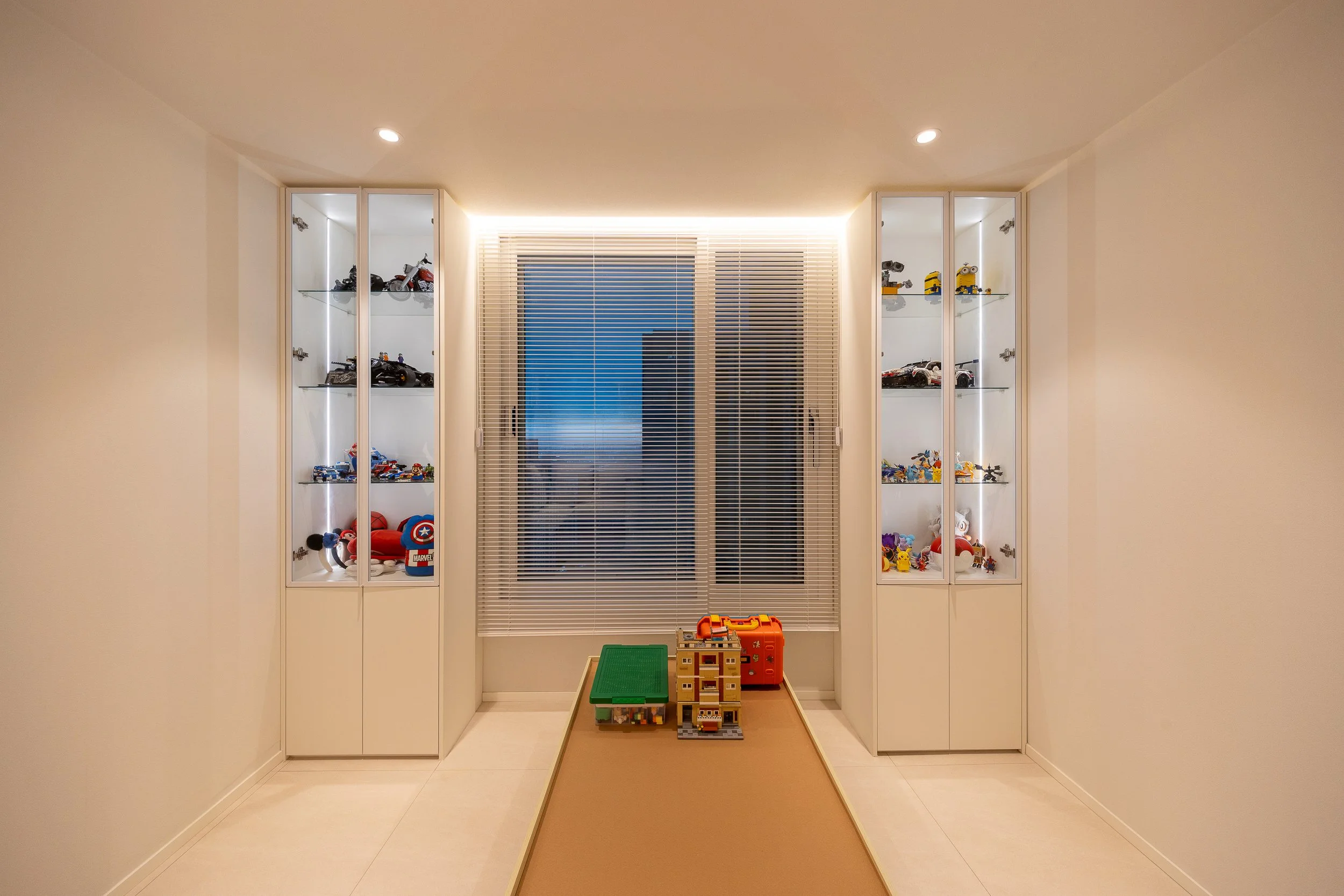 A room with two glass display cabinets filled with toy cars and action figures, a window with blinds, and a narrow table with toys in the center.