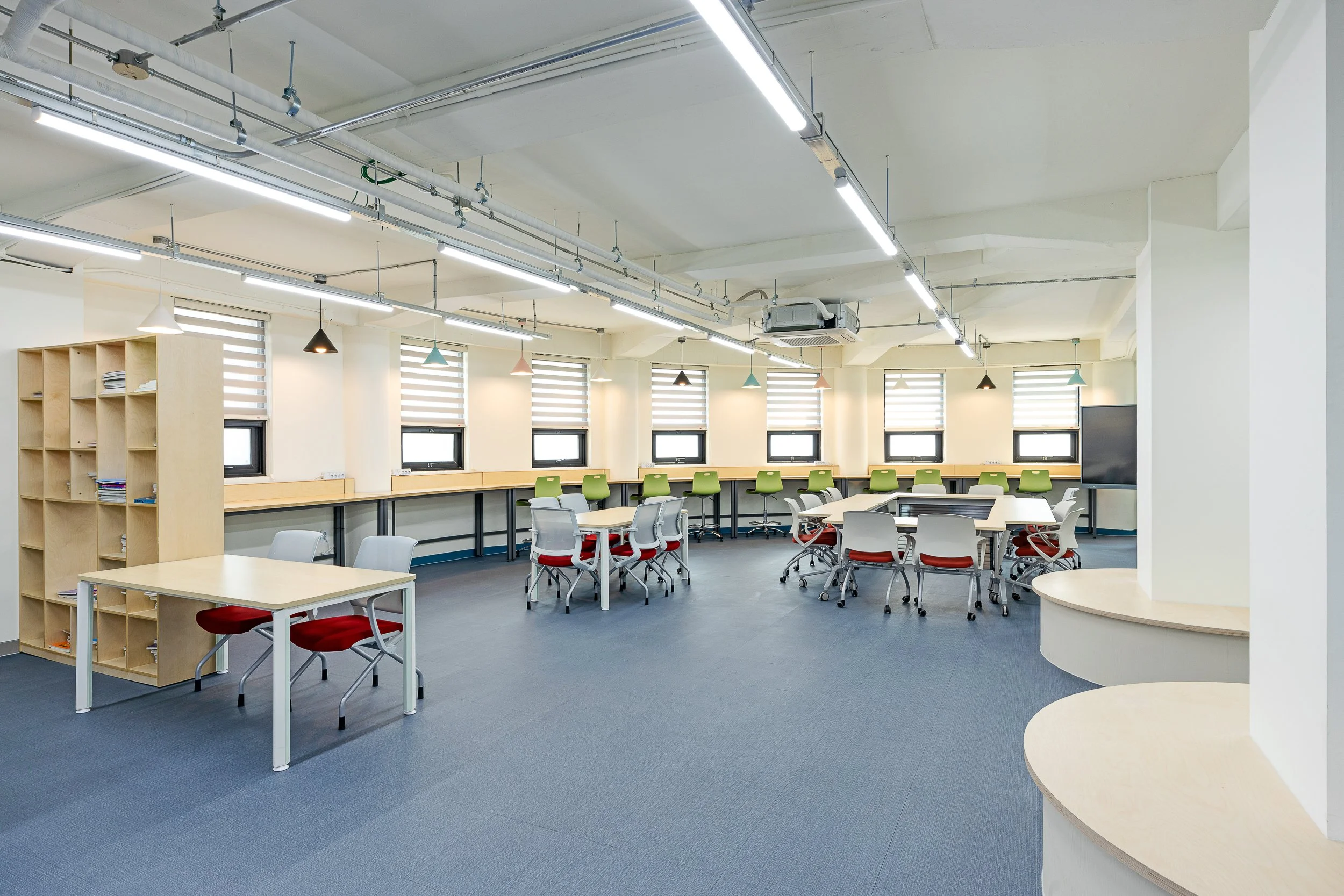 Empty classroom with multiple desks and chairs, windows with blinds, and modern lighting fixtures.