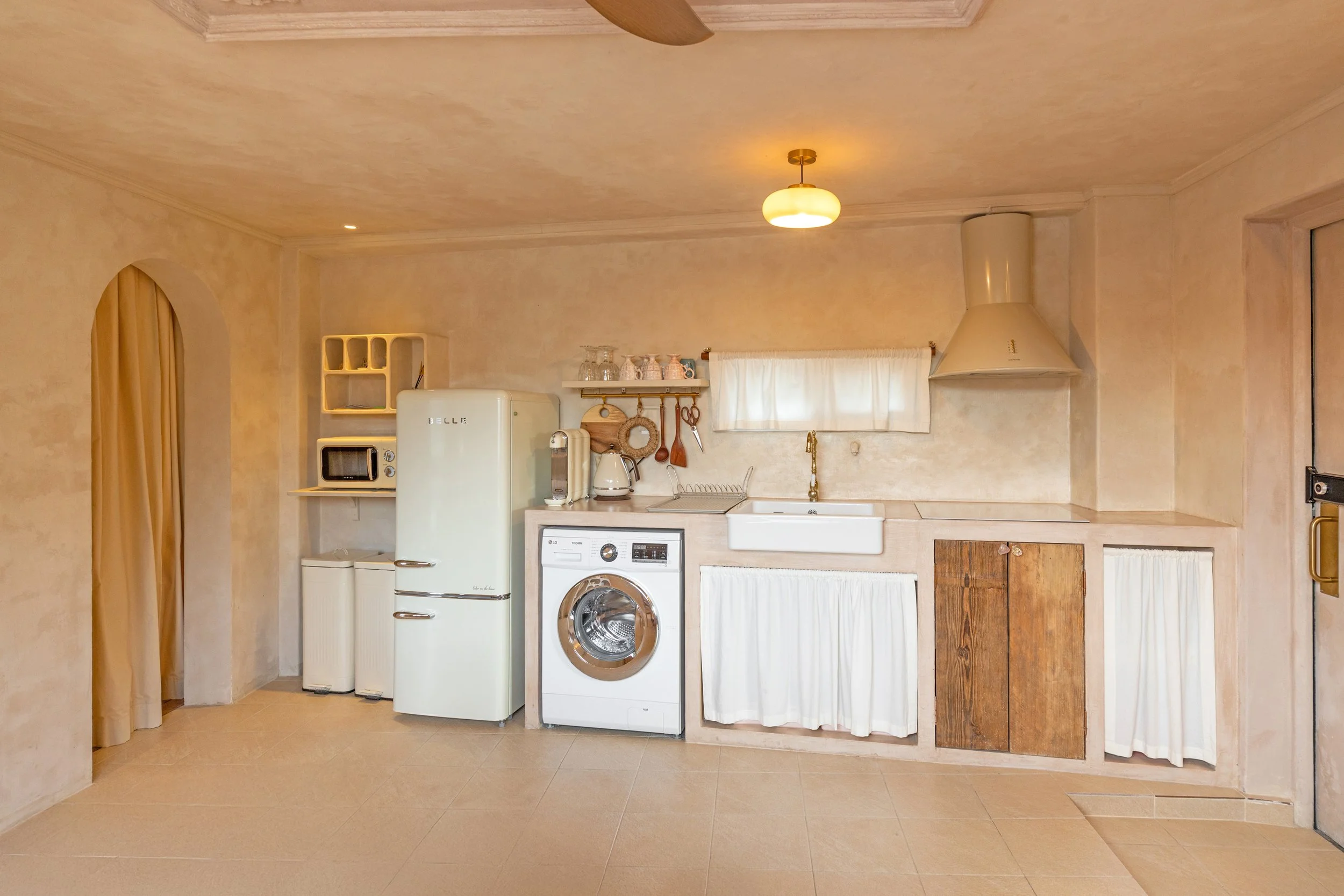 A vintage-style kitchen with beige walls, a white refrigerator, a microwave, a washing machine, a small window with a curtain, and wooden cabinets covered with white curtains. There is a white farmhouse sink with a gold faucet, and a small shelf with