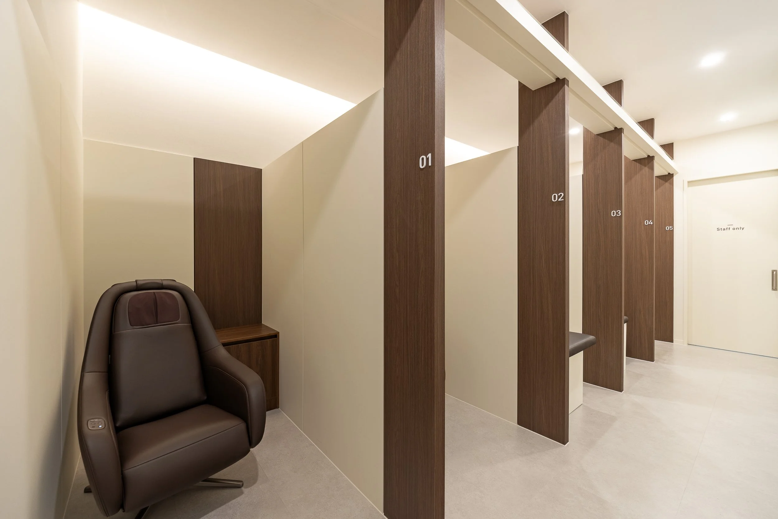 A row of individual privacy stalls with numbered wooden partitions in a modern, well-lit lounge or spa area. An empty black massage chair is positioned outside the stalls.