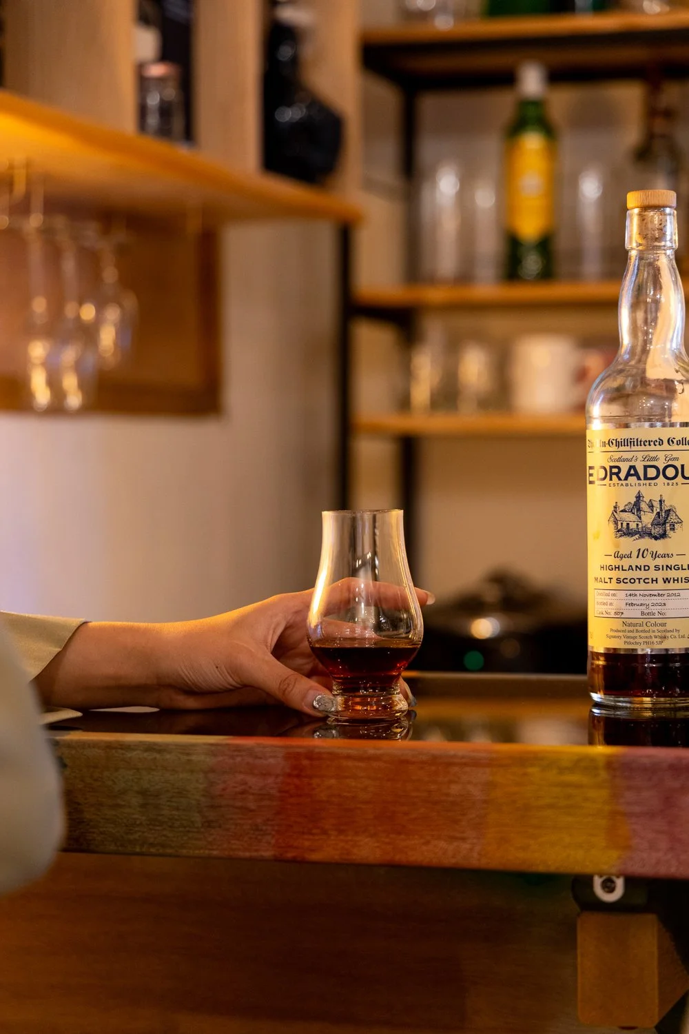 Person holding a glass of dark whiskey at a bar with a bottle of GlenOrd Scotch whiskey on the counter.