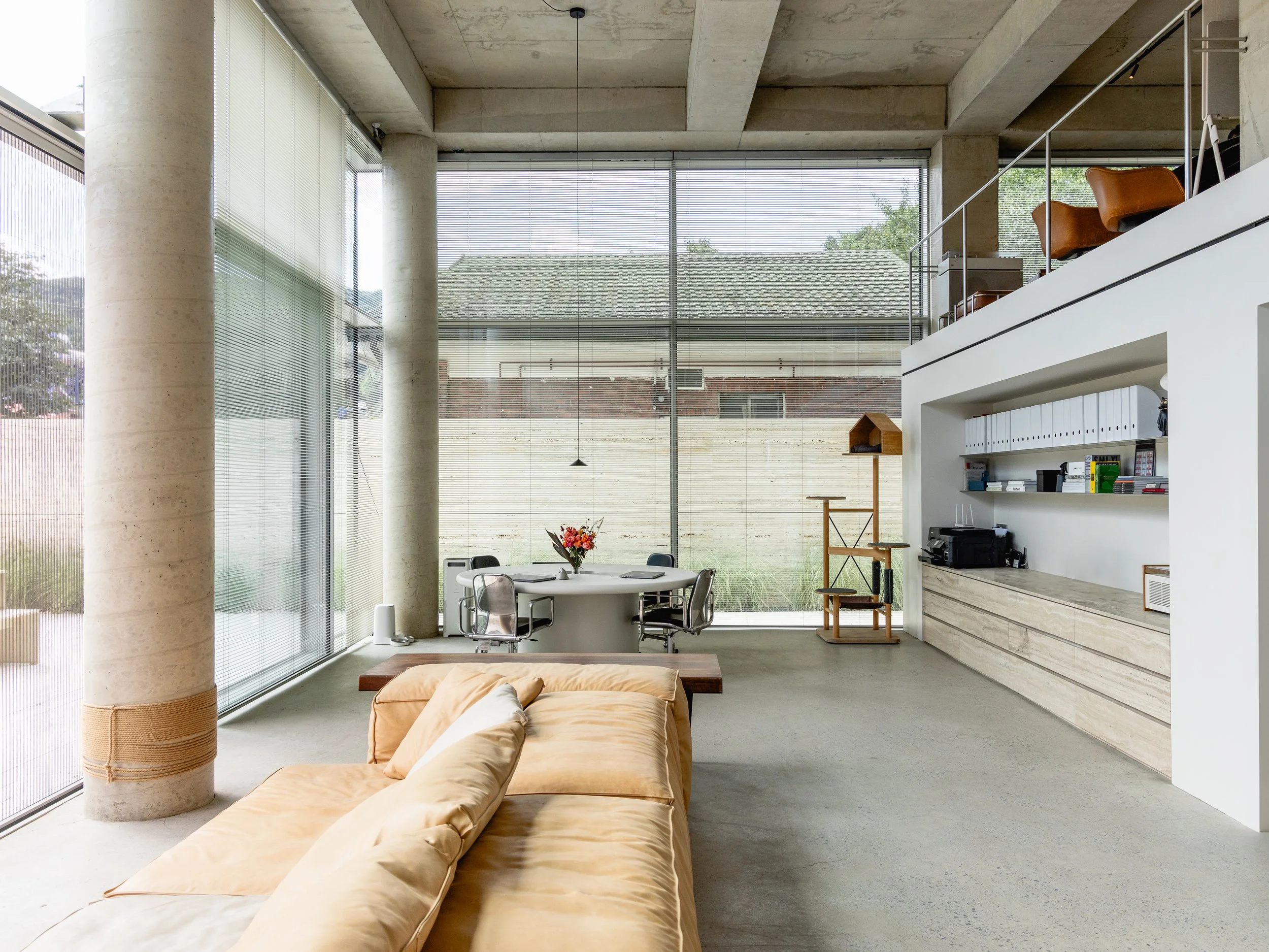 Modern living room with large floor-to-ceiling windows, a beige sofa, a round dining table with chairs, and a high ceiling with concrete beams.